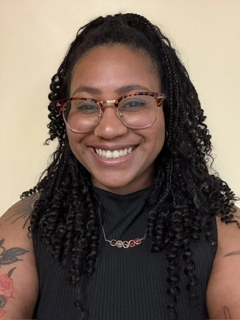 A woman with curly black hair in braids, wearing glasses with tortoiseshell frames, smiling, dressed in a sleeveless black top, with a tattoo on her right arm, and a necklace with small red and gold elements.