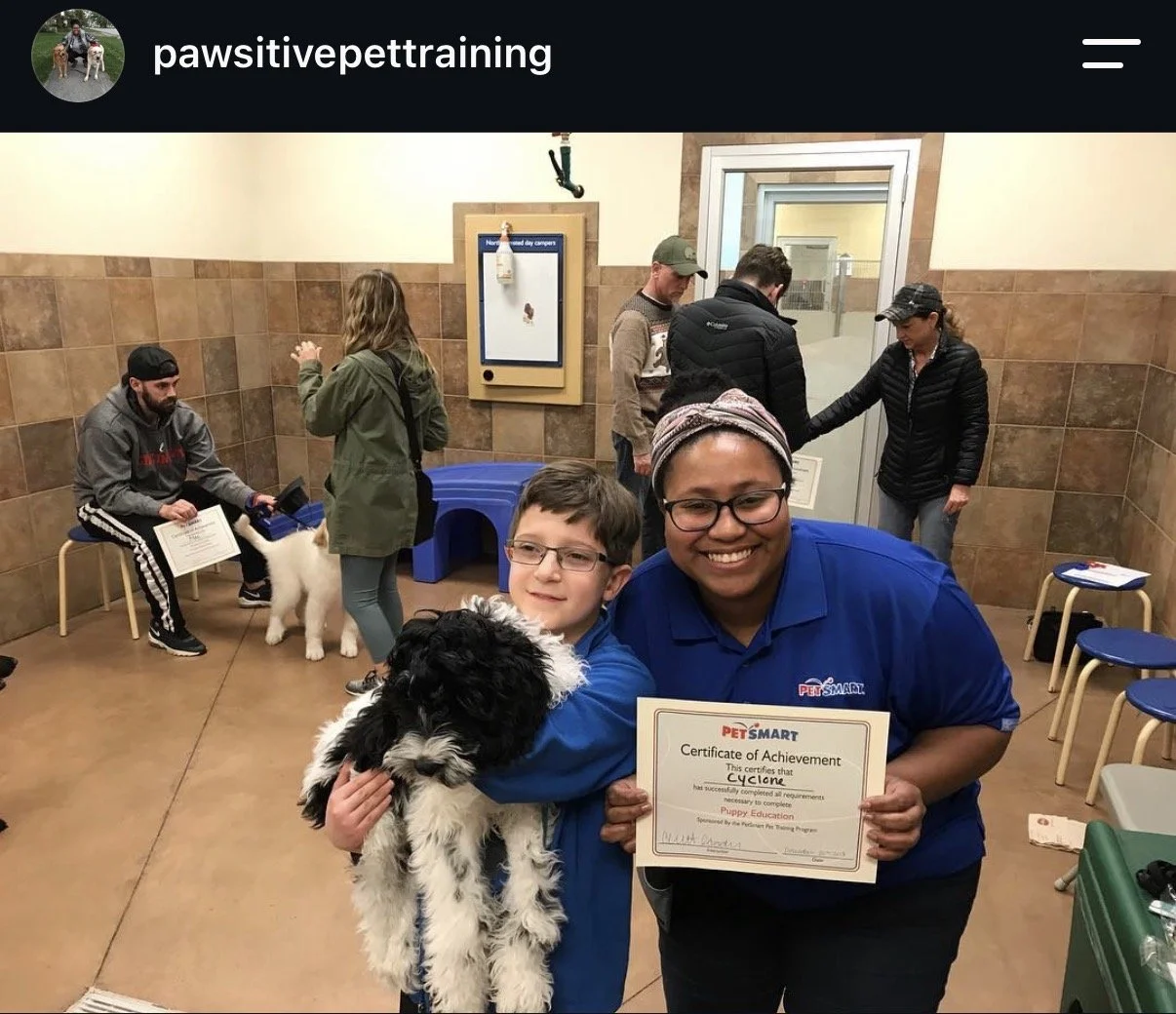 A young boy holding a black and white puppy is standing next to a smiling woman in a blue PetSmart uniform, who is holding a certificate of achievement for puppy training. In the background, several people are gathered, some with dogs, inside a PetSmart store.