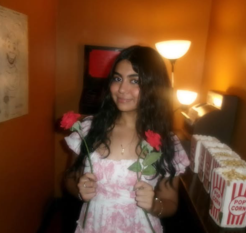 Young woman with long dark curly hair holding two red carnations, standing in a warmly lit room with orange walls, a lamp, and a box of popcorn.