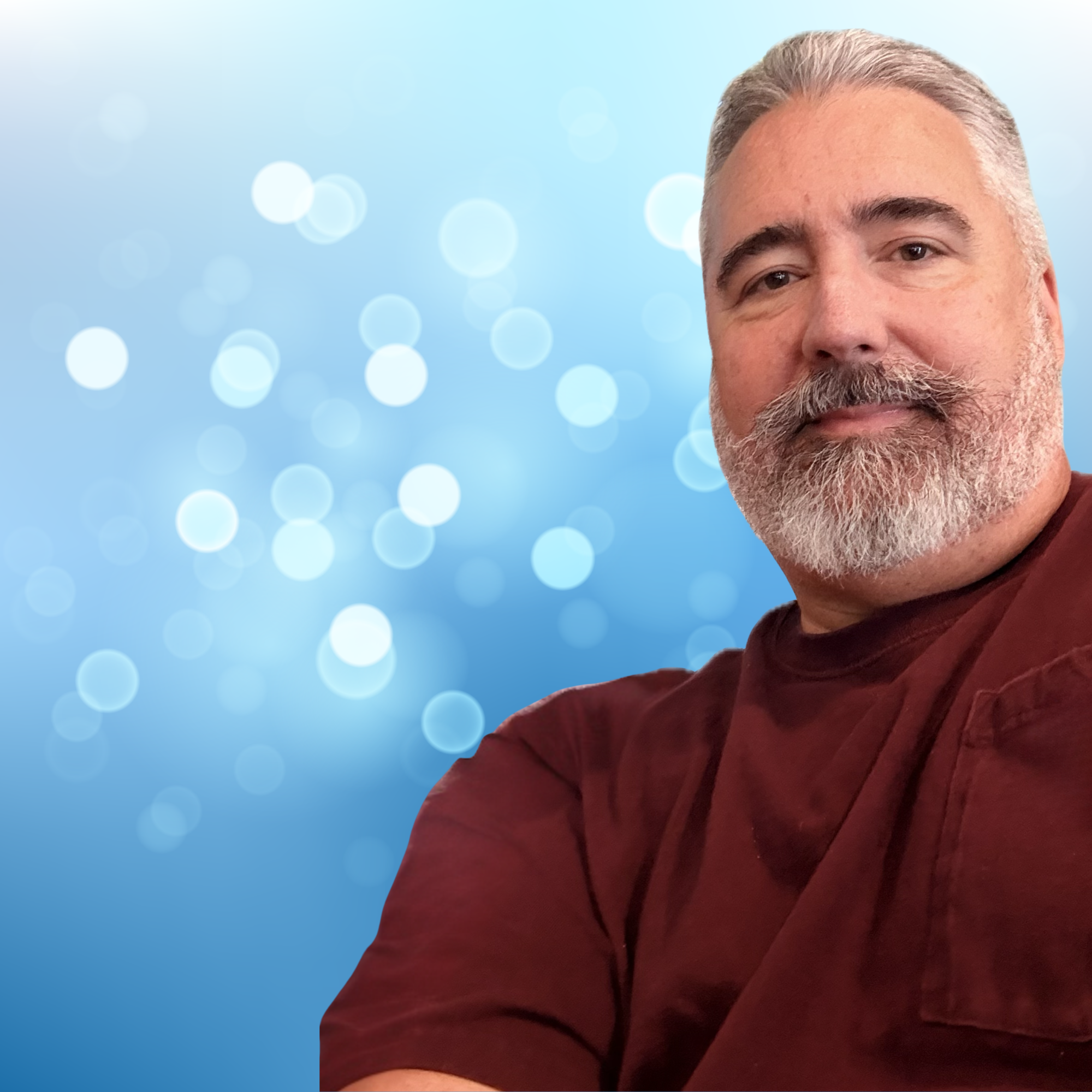 Close-up of a middle-aged man with gray hair and a beard, smiling slightly, wearing a maroon shirt or sweater, with a blue bokeh background.