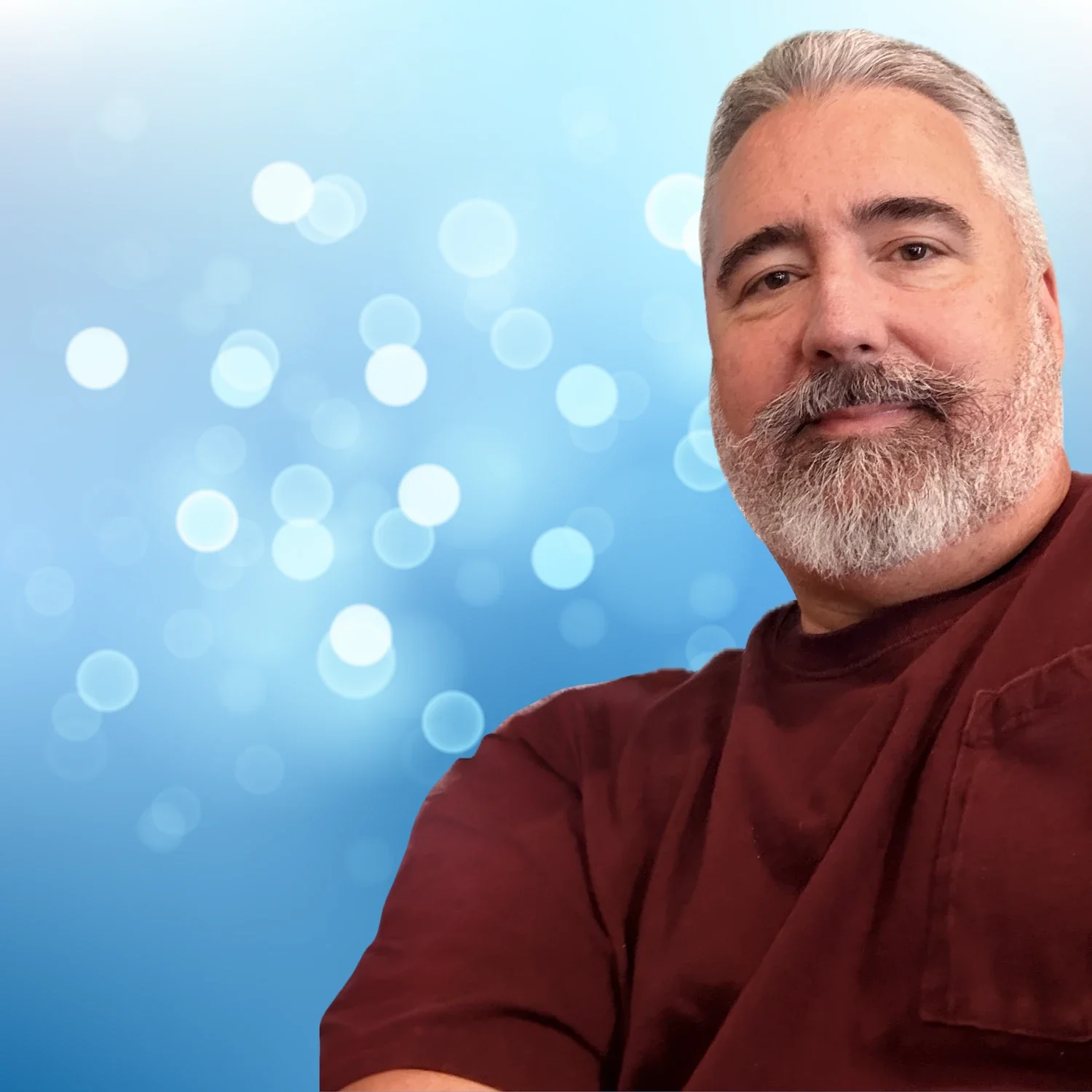 A man with gray hair and a beard taking a selfie against a light blue bokeh background.