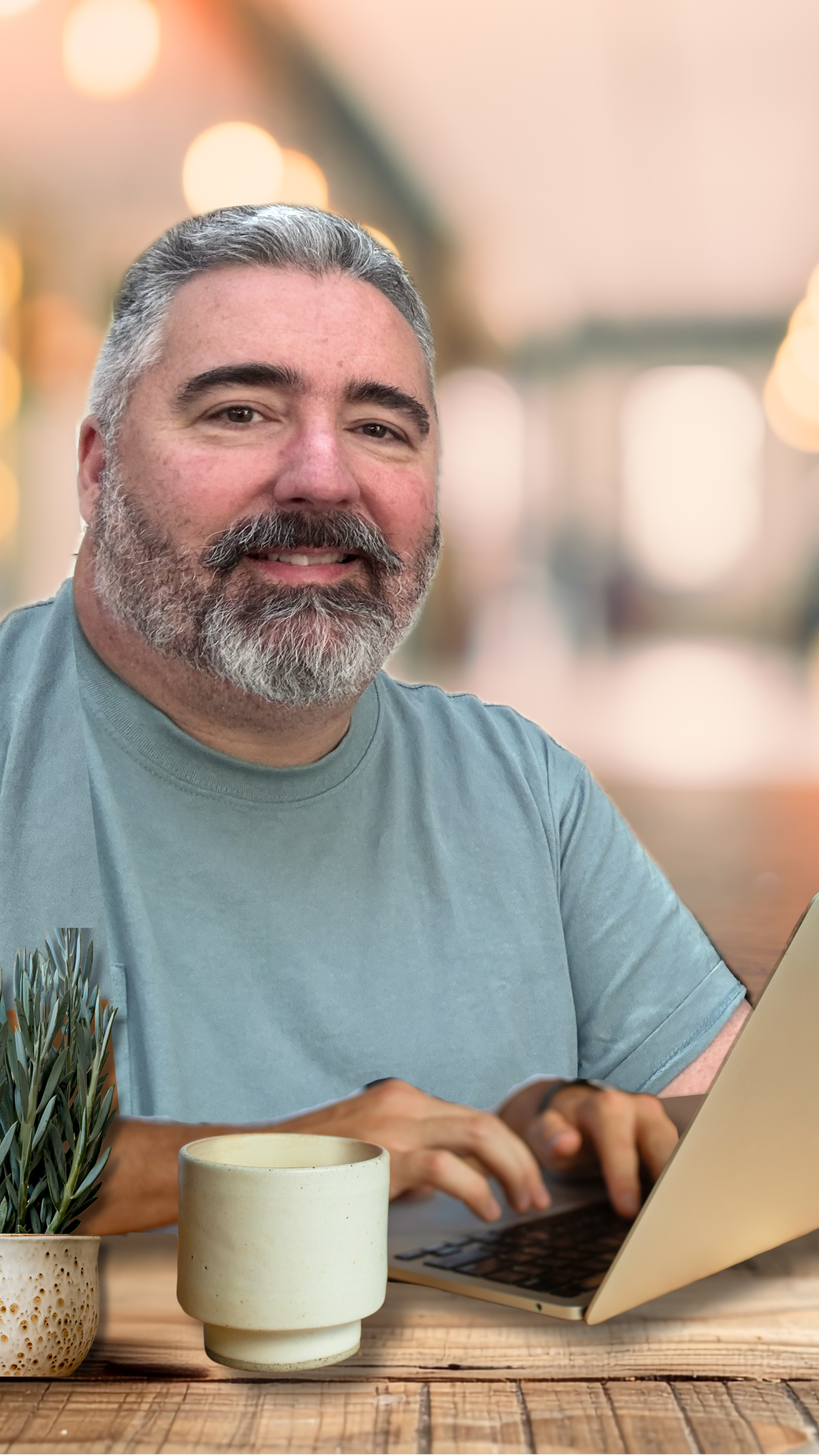 A man with gray hair and beard working on a laptop at a wooden table, with plants and a coffee mug in the foreground, in a warmly lit indoor setting.