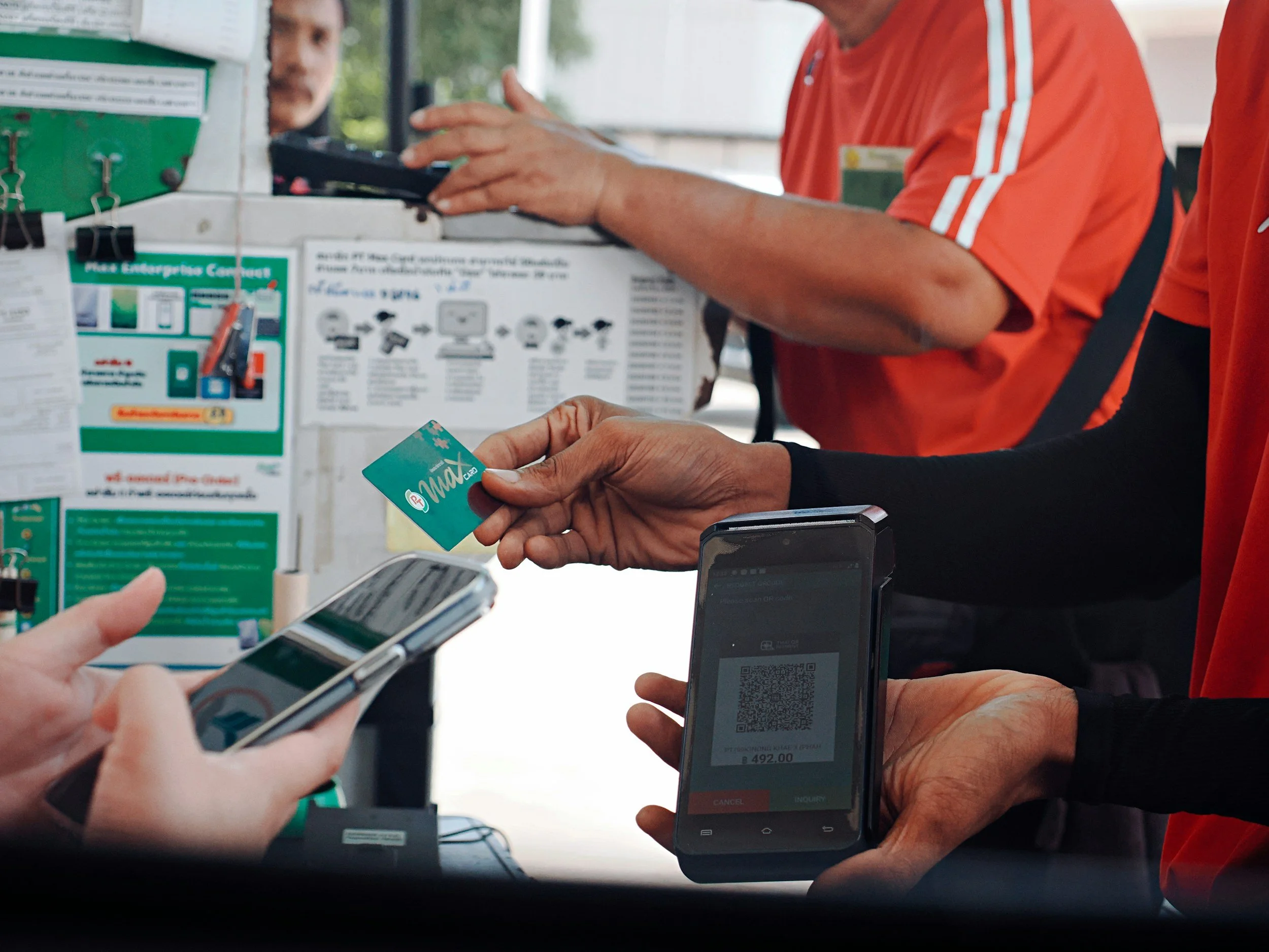 People using smartphones to scan QR codes at a checkout counter, with a cashier in orange shirt assisting.