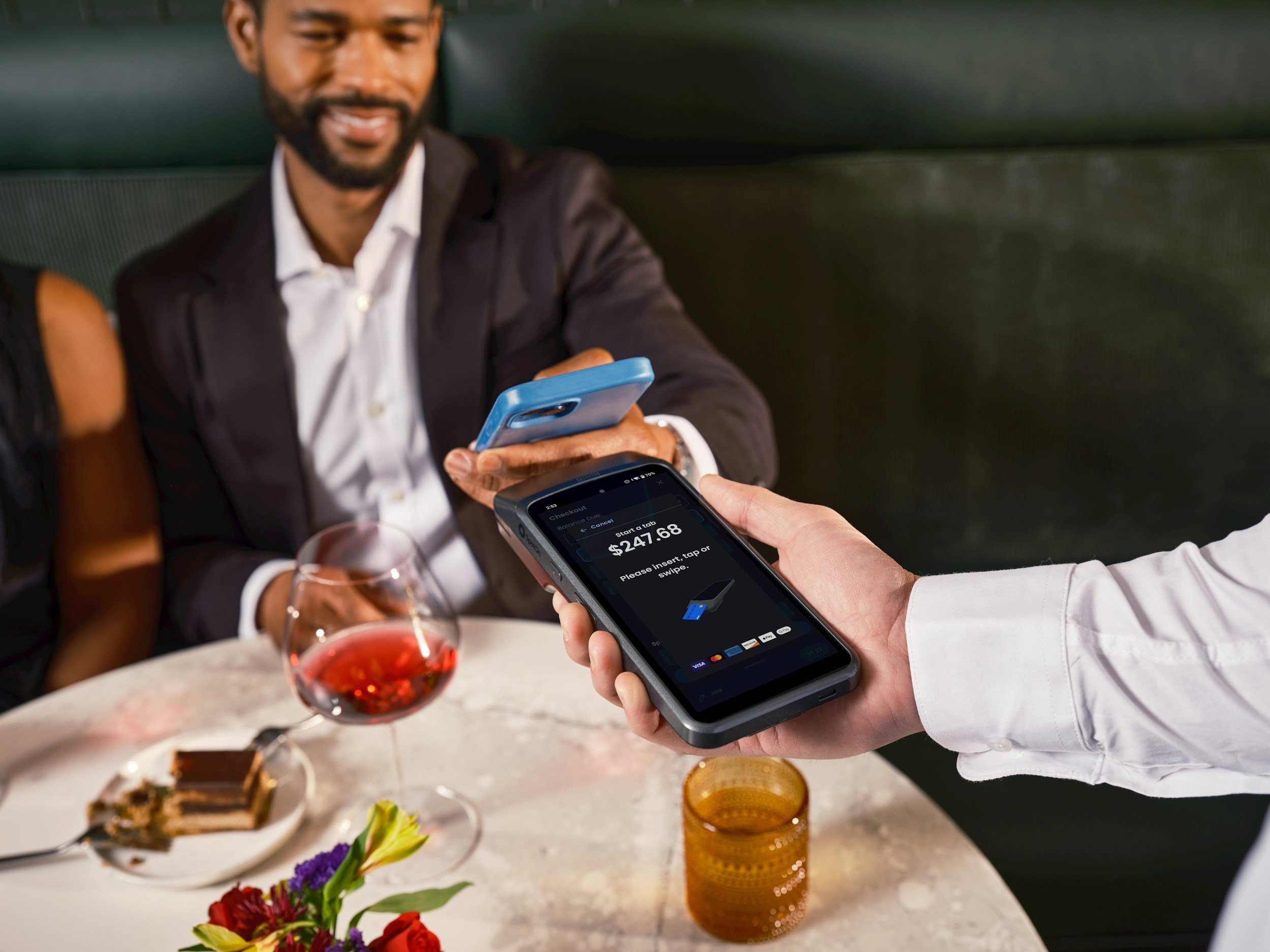 A man in a suit and white shirt is smiling as he uses a contactless payment terminal, with a woman holding a smartphone nearby. On the table, there is a glass of red wine, a plate with dessert, and a vase with flowers.