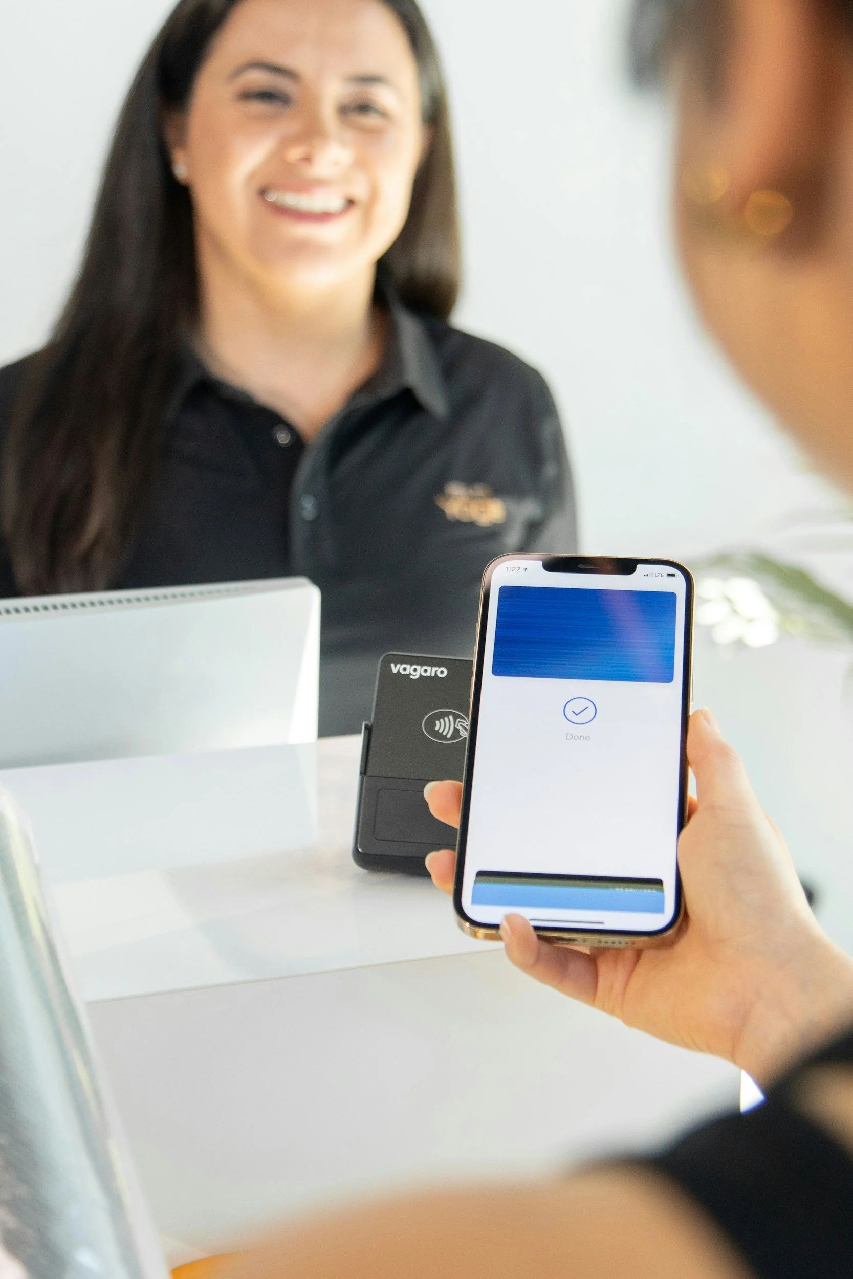 A woman in a black shirt at a counter, with a Galaxy device and another person's hand holding a smartphone displaying a confirmation screen, in an indoor setting.