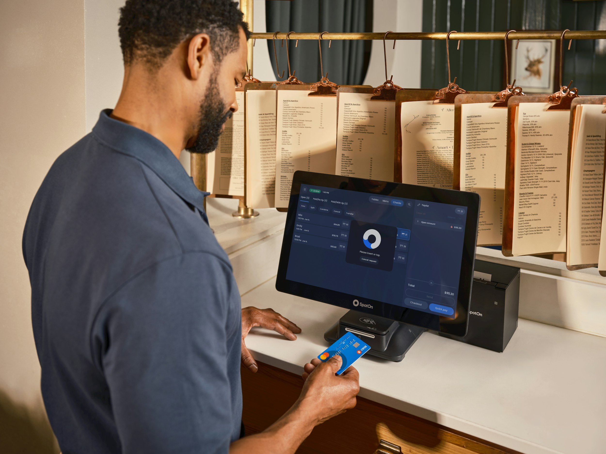 A man with short curly hair and a beard is making a payment using a credit card at a checkout kiosk in a store. He holds a blue credit card in his right hand, and the electronic payment terminal is placed on the counter in front of him. The man is wearing a dark blue shirt, and the background features a gold-colored metal rack with multiple menus or signs hanging from it.