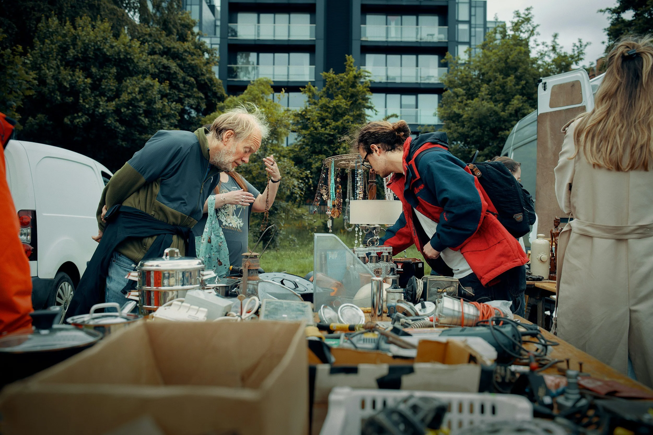 People browsing vintage and secondhand items at an outdoor market with a modern building and trees in the background.