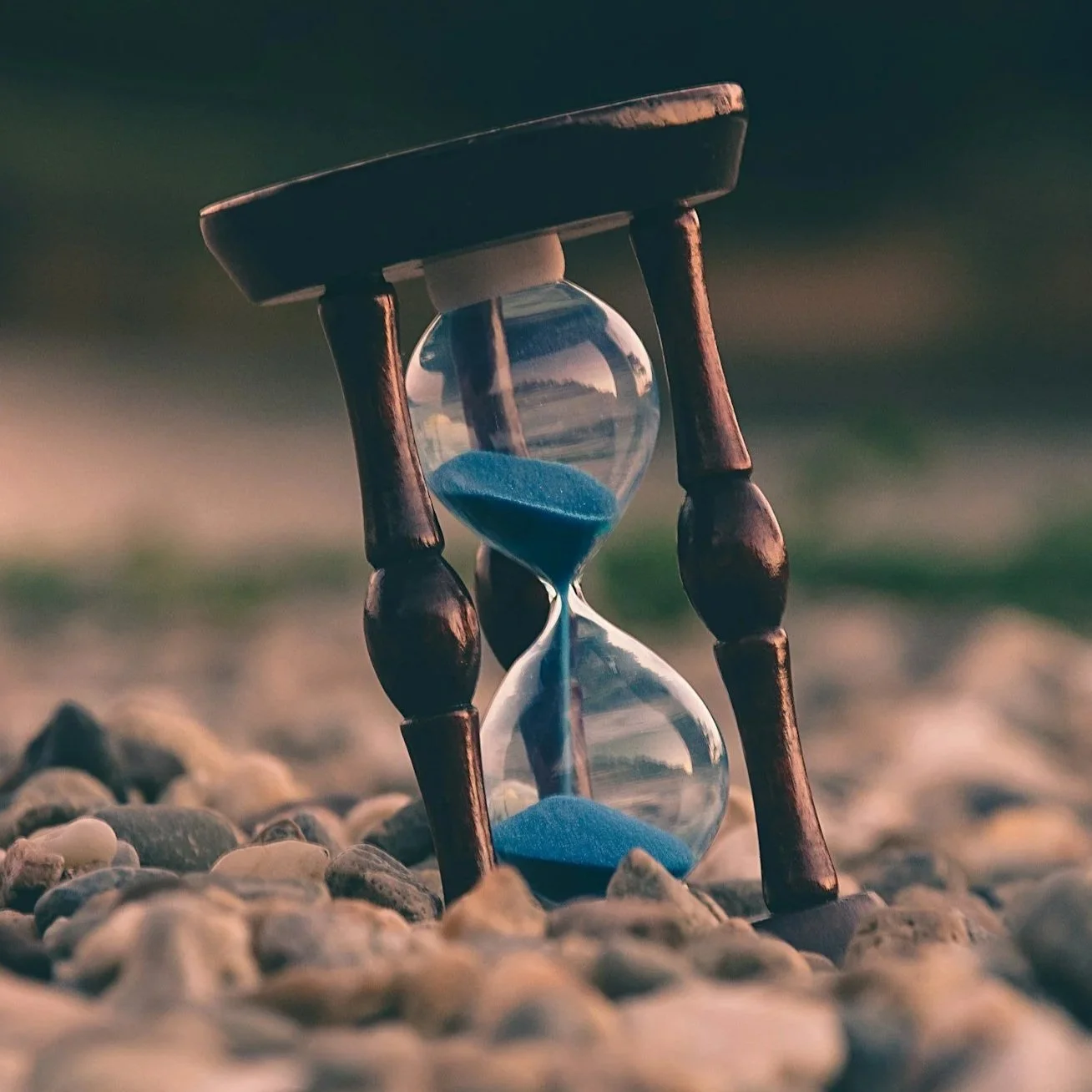 An hourglass with blue sand standing on a bed of small pebbles outdoors.