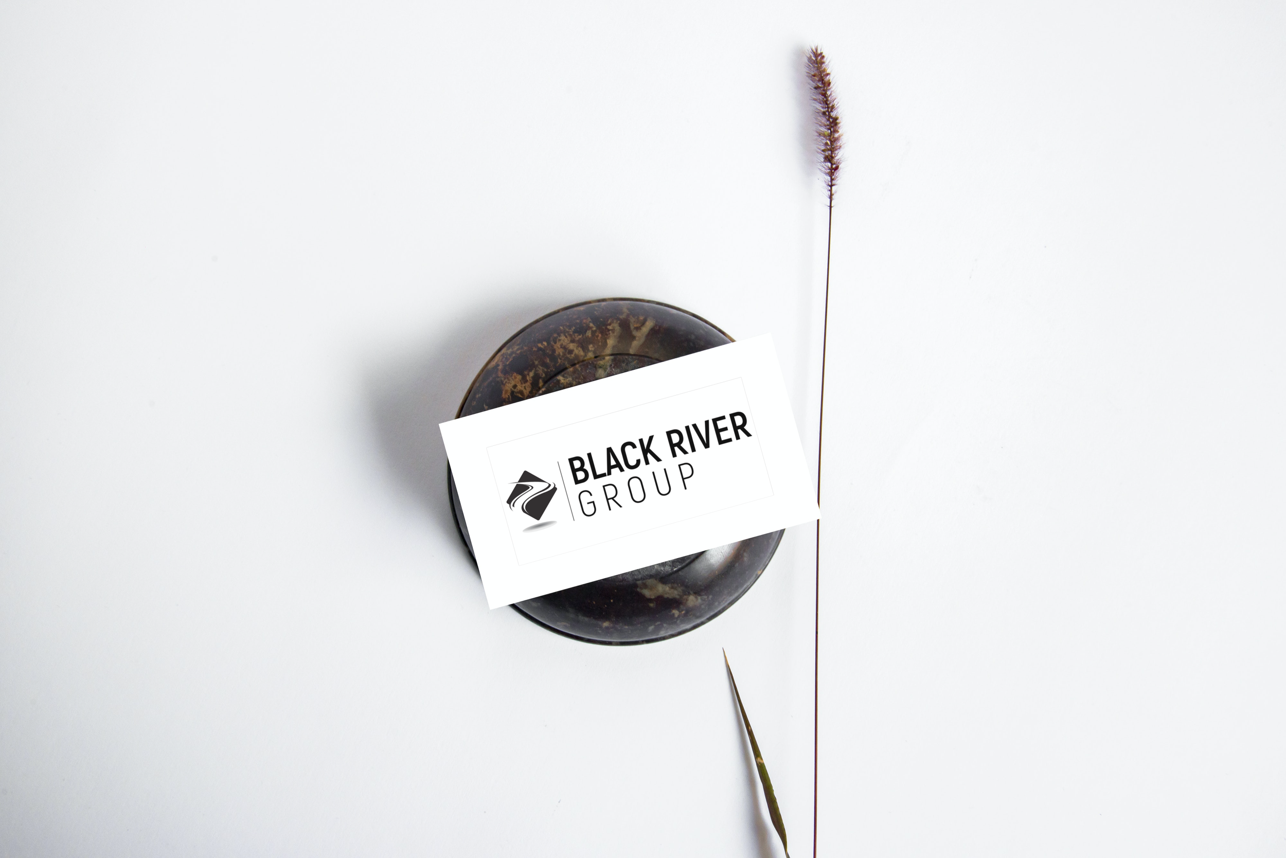 A round dark stone with a thin stick or plant stem on top of it, and a business card with the logo and text 'Black River Group' placed partially on the stone. The background is plain white.