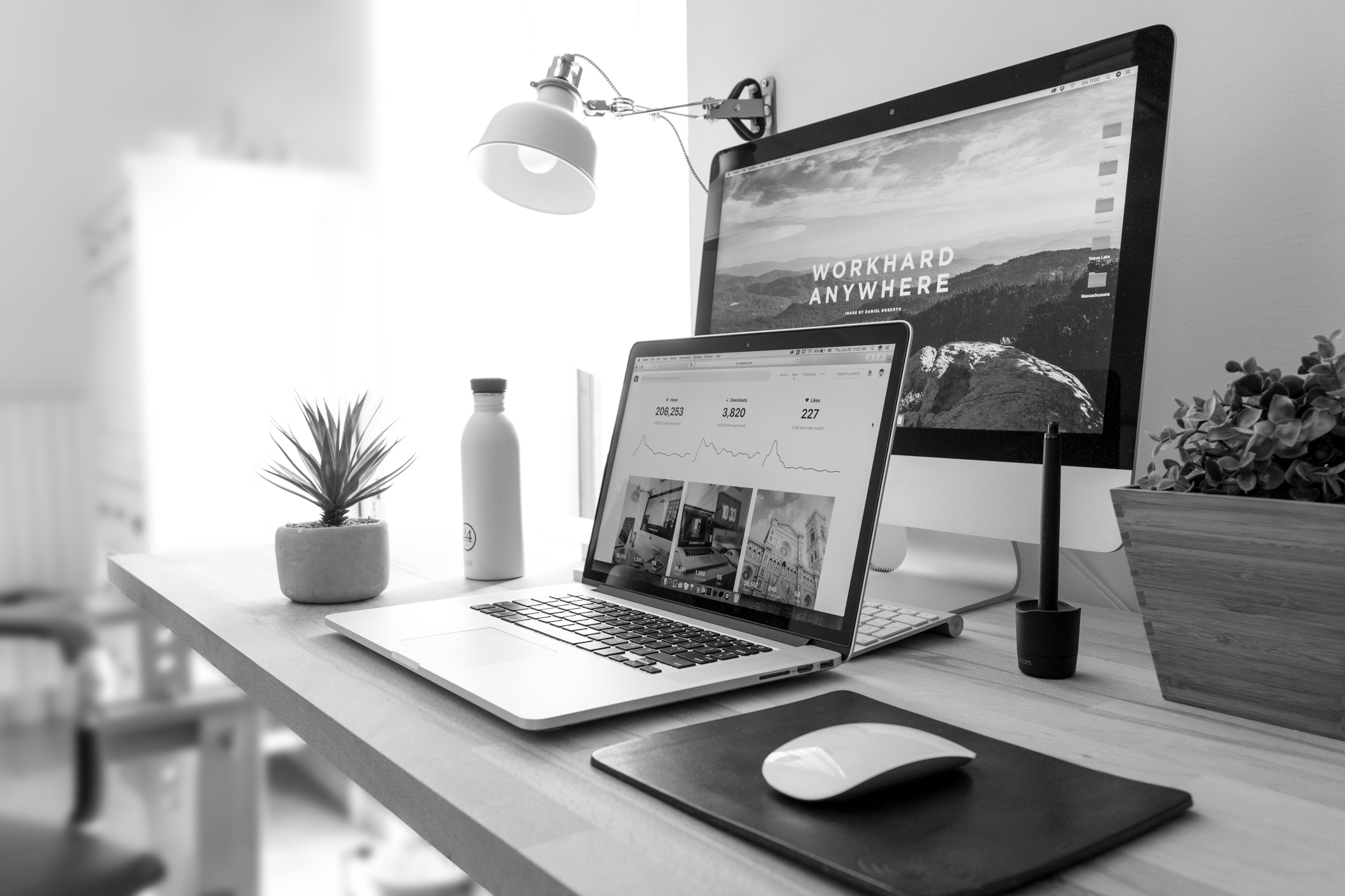 A desk workspace with a MacBook laptop displaying images and a separate desktop monitor showing a mountain landscape with the text "Workhard Anywhere." The desk has a small potted succulent plant, a water bottle, and a white computer mouse. There is a wall-mounted adjustable lamp above the monitor, and a black cup or container holding pens is next to the monitor.
