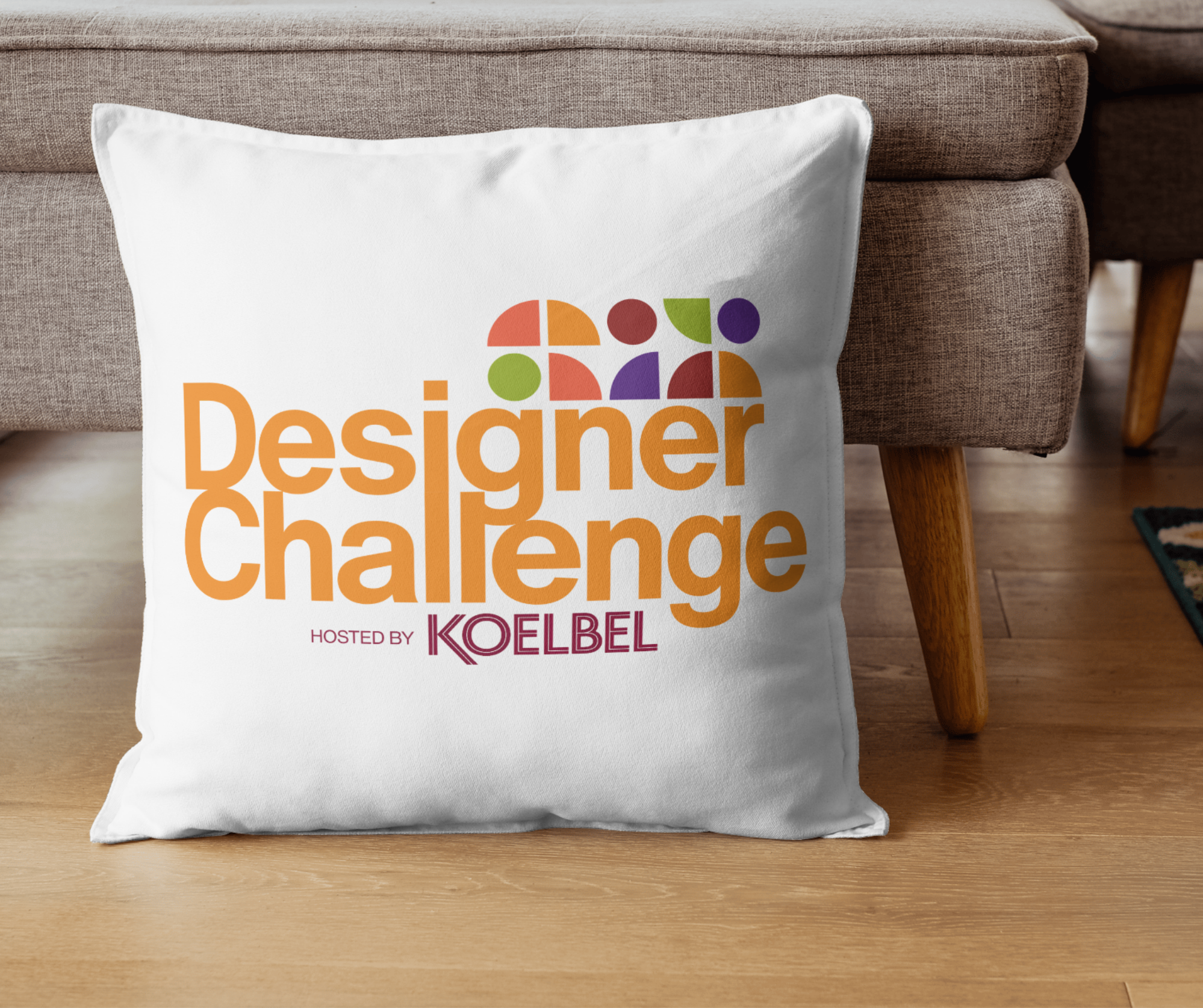 A white cushion with colorful text and design elements resting on a wooden floor next to a beige sofa with wooden legs.