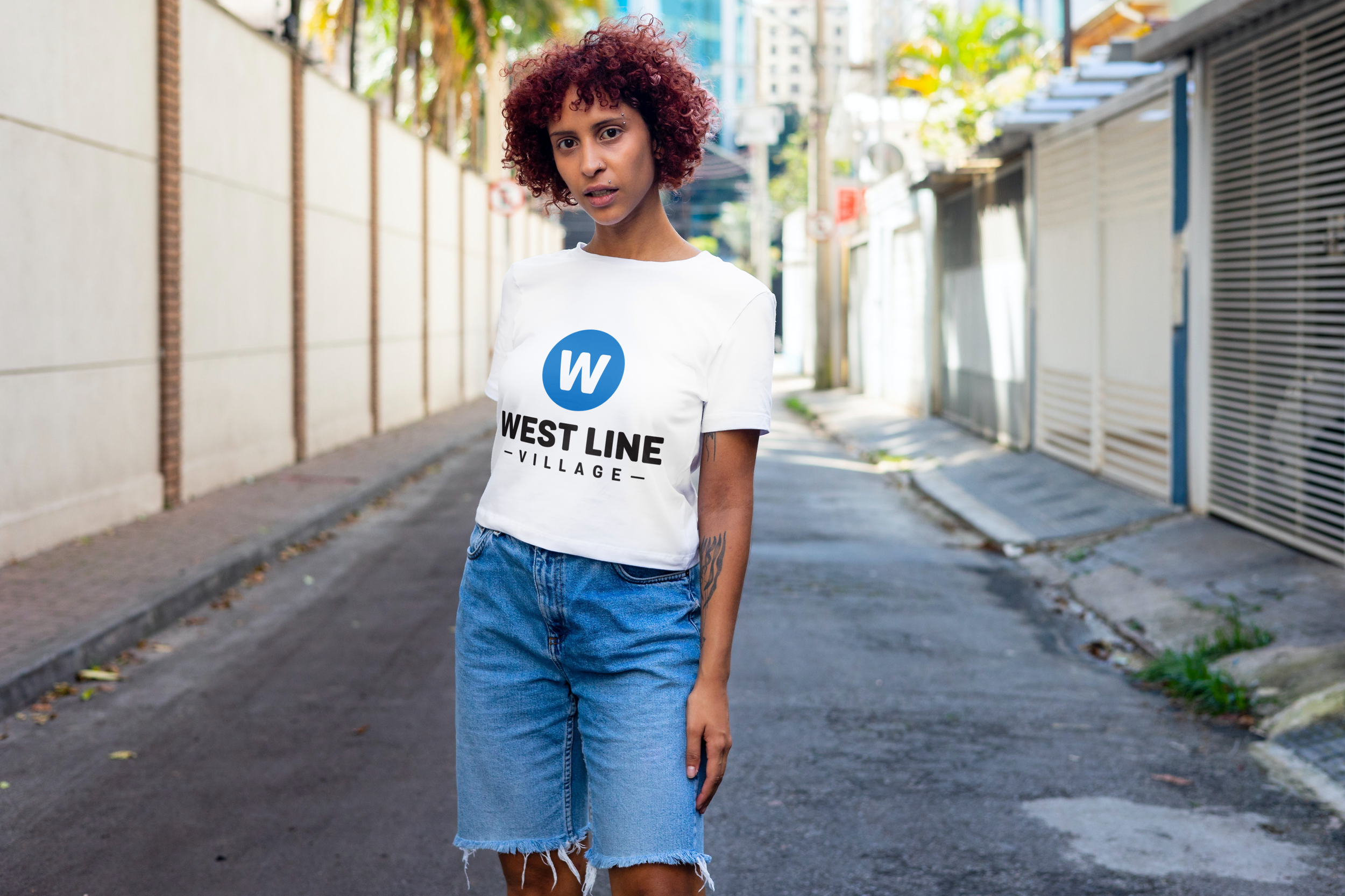 A woman with curly hair wearing a white T-shirt with the words 'WEST LINE VILLAGE' and a blue 'W' logo, standing on a city street.