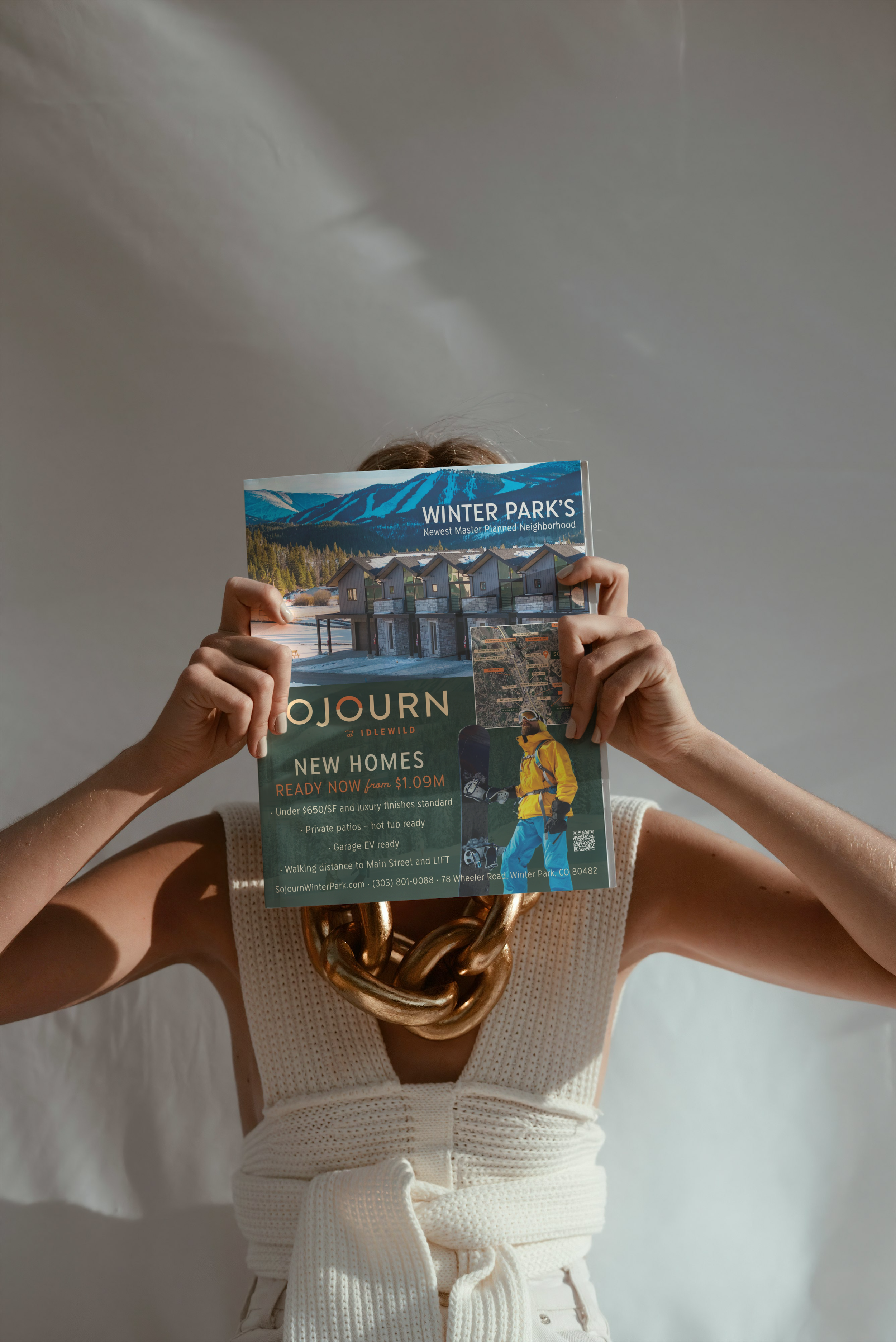 Person wearing a large gold chain necklace and white knitted top, holding and covering their face with a colorful flyer for Winter Park homes and neighborhoods.