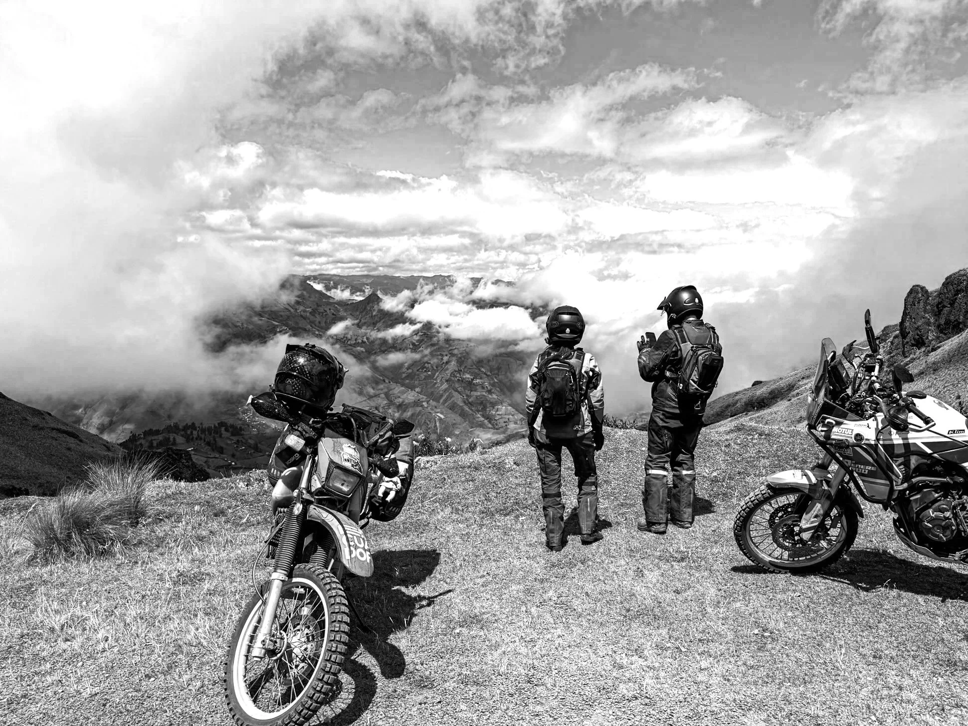 Three motorcyclists in protective gear and helmets standing on a mountain overlook, with two off-road motorcycles parked nearby and a scenic view of mountains and clouds in the background.