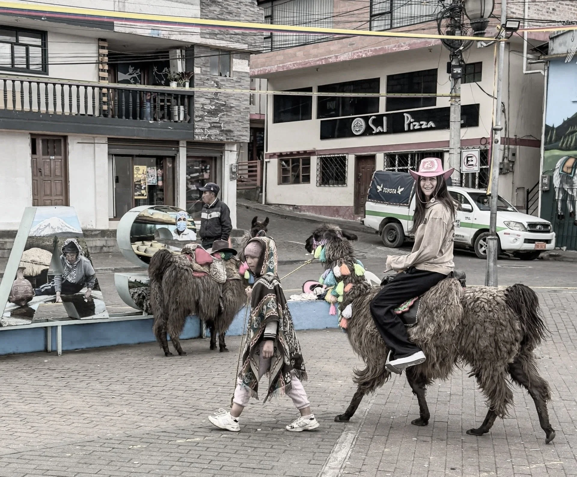 A young woman wearing a pink cowboy hat and beige jacket riding a llama. A girl with a patterned coat walks nearby. Several llamas with colorful pom-pom accessories are in the scene. In the background, there are people, parked cars, and storefronts, including one with a sign that says 'Sal Pizza.'