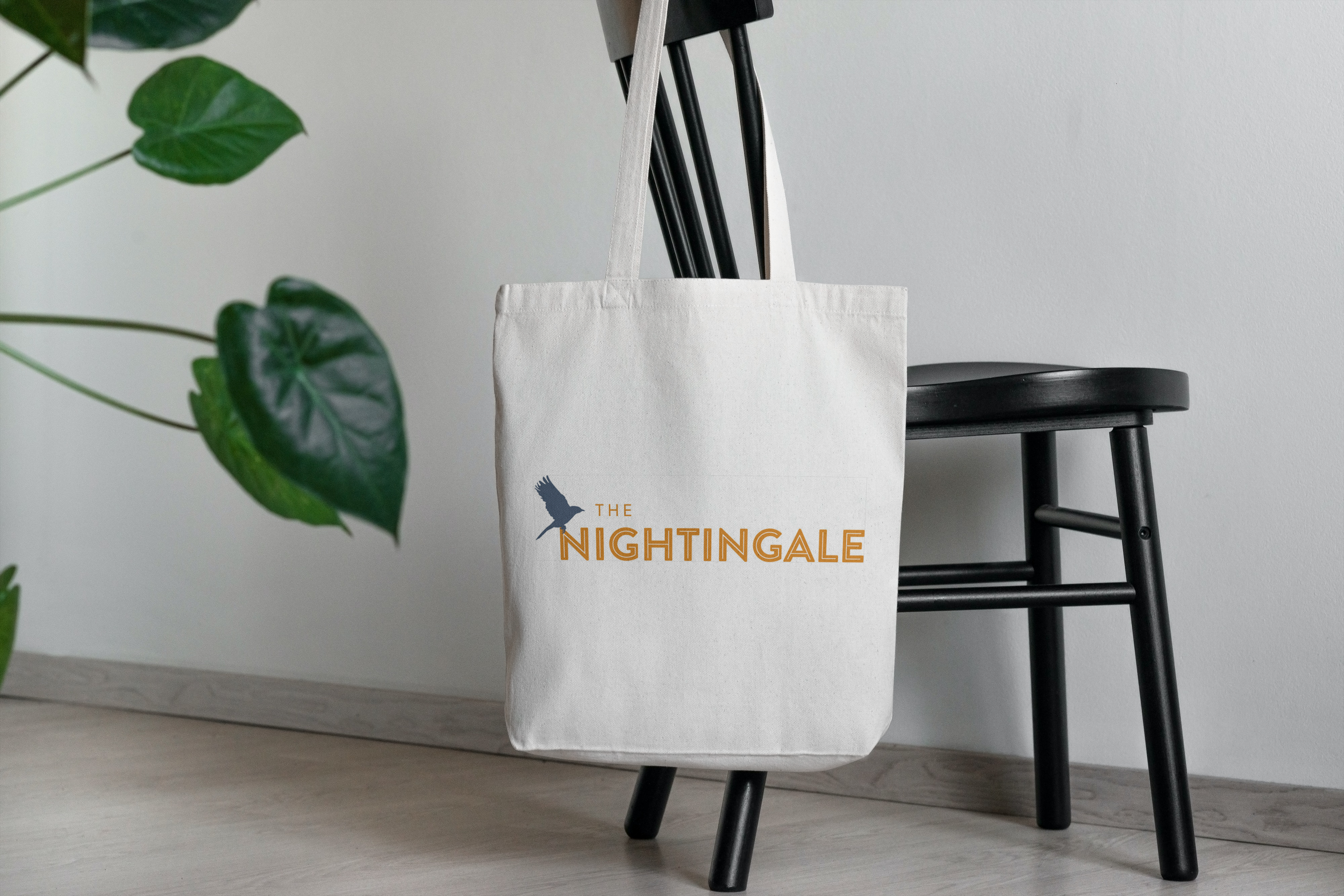 A white tote bag with orange text reading 'The Nightingale' and a silhouette of a bird, hanging on the back of a black wooden chair next to a white wall and green plant leaves.