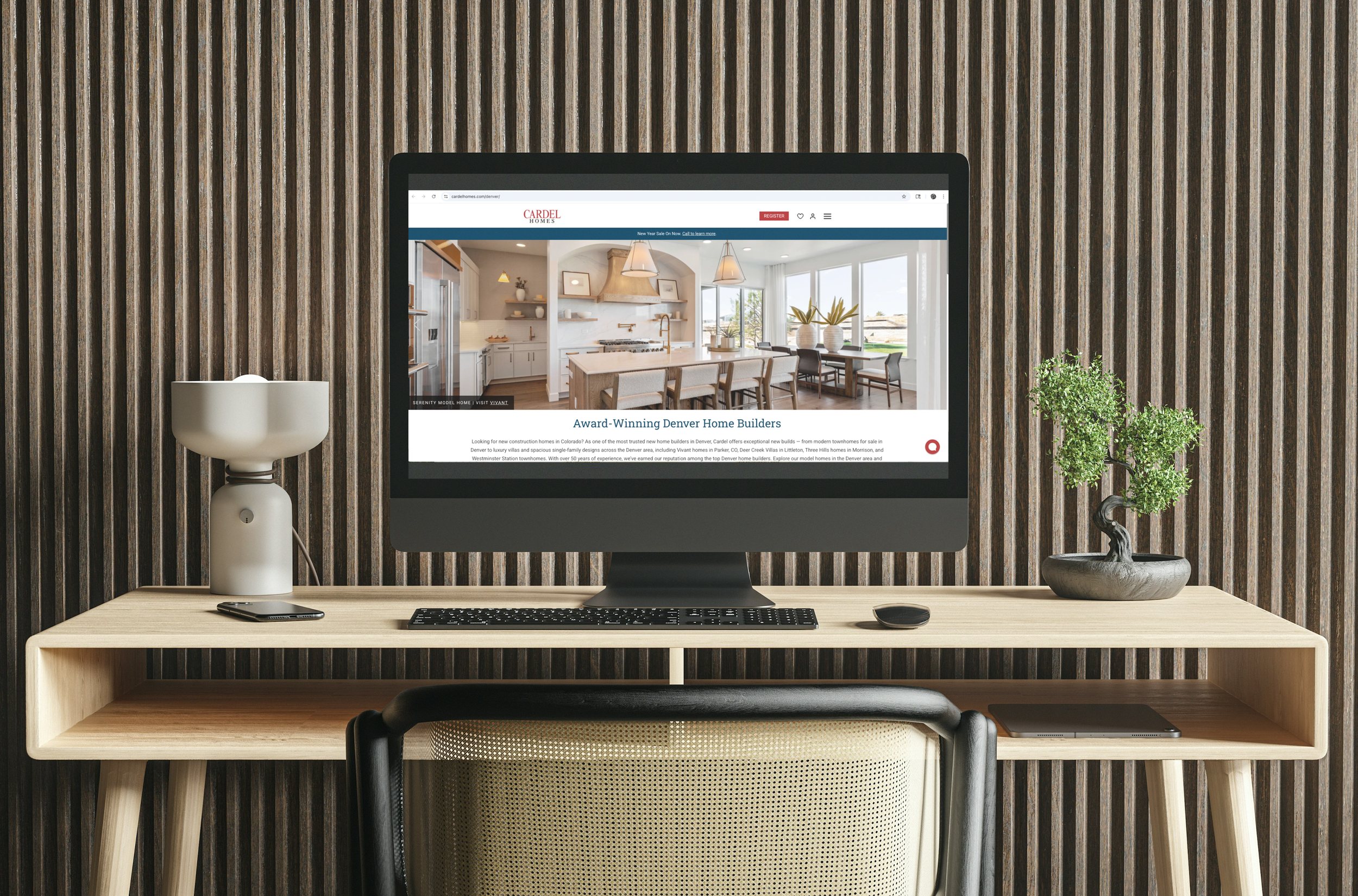 Computer monitor displaying a real estate website with images of a modern kitchen and dining area, on a wooden desk flanked by plants and a lamp, against a vertical wooden slat wall.