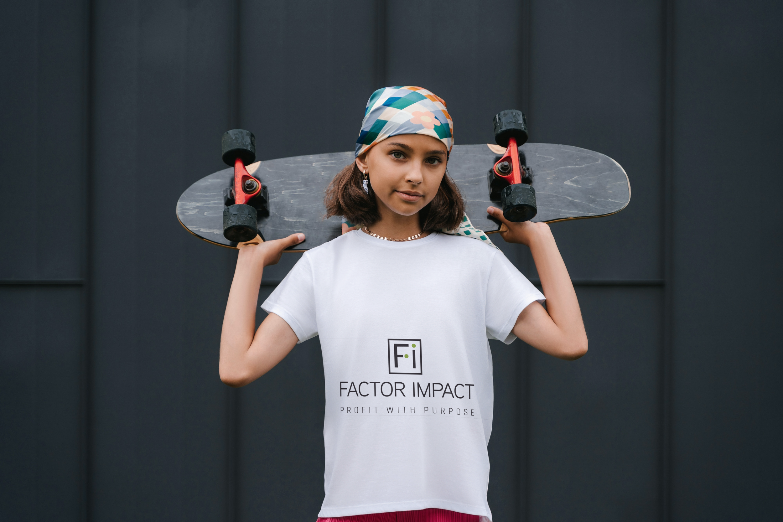 Young woman with brown hair wearing a colorful headscarf, earrings, and a white t-shirt with the text 'Factor Impact' and holding a skateboard on her shoulders, standing against a gray wall.