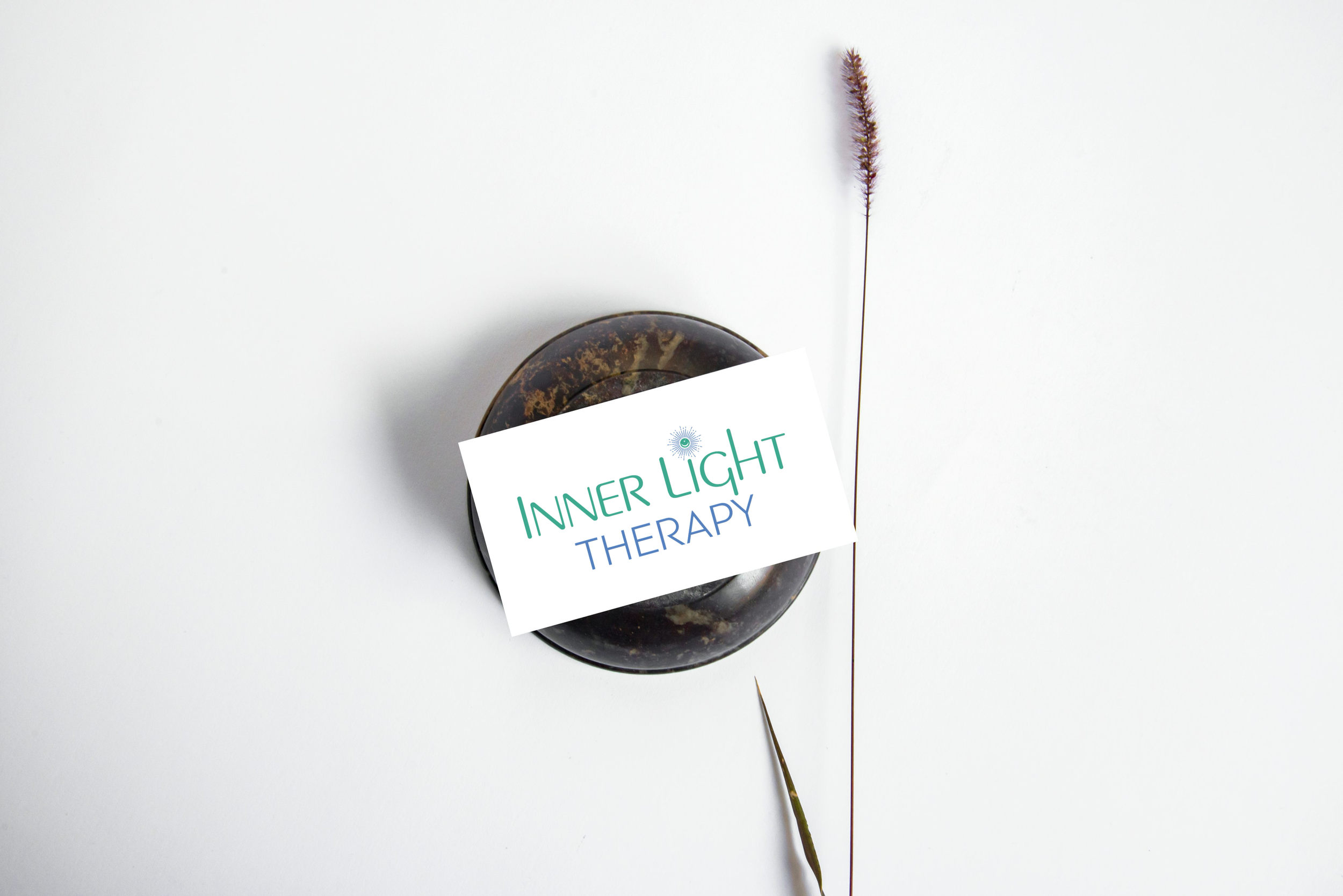 A round dark stone with a sign on top that reads "Inner Light Therapy." There is a tall, thin grass seed head to the right of the stone, and a white background with a faint shadow of the grass seed head.
