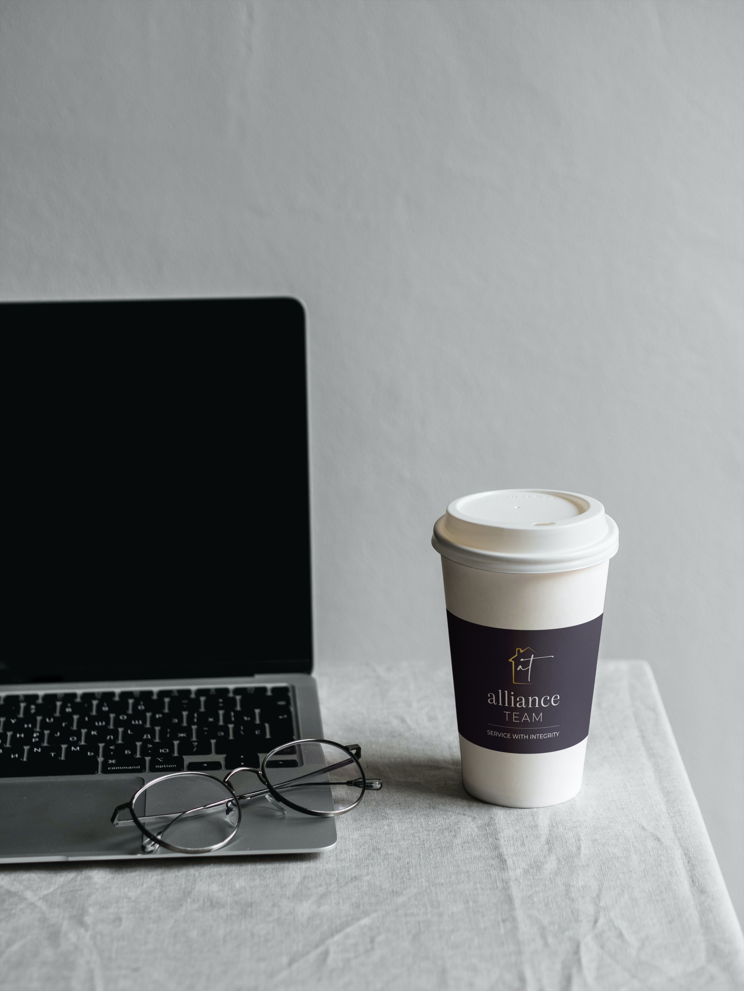 A workspace with a partially visible laptop, a pair of eyeglasses, a cup of coffee with a sleeve labeled 'alliance TEAM,' and a pair of eyeglasses on a desk.
