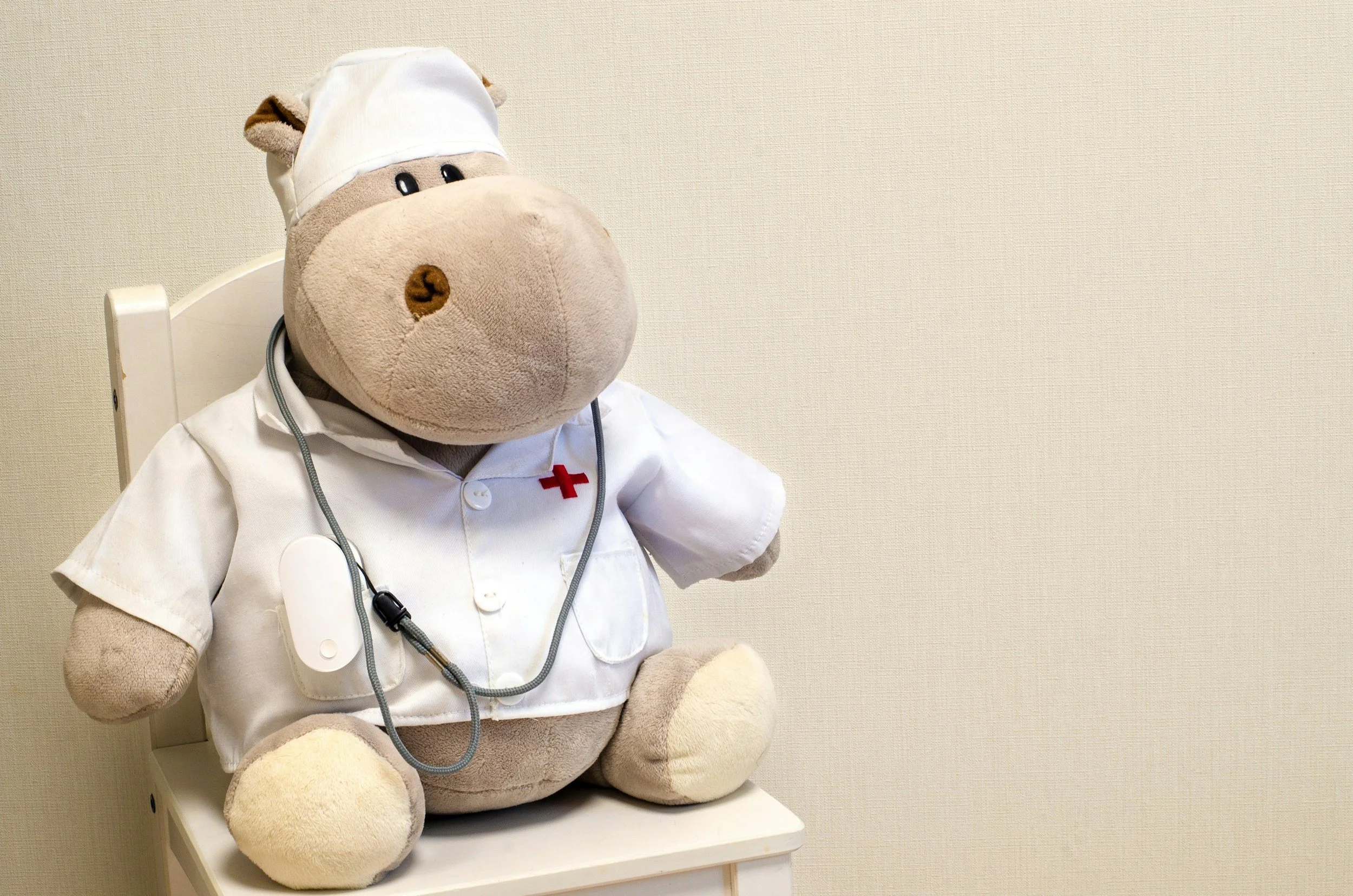A plush teddy bear dressed as a nurse, sitting on a medical examination table against a plain wall. The bear wears a white nurse's hat with a red cross, a white coat, and has a stethoscope around its neck.