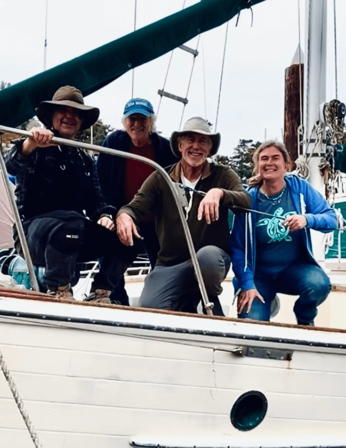 Four people on a sailboat smiling and posing for a photo, wearing outdoor clothing and hats, with a cloudy sky in the background.