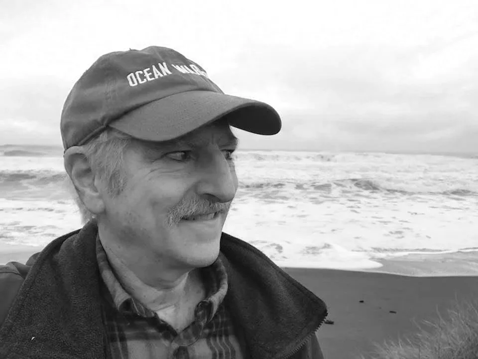 David Helvarg wearing a cap and jacket standing on a beach with ocean waves in the background.