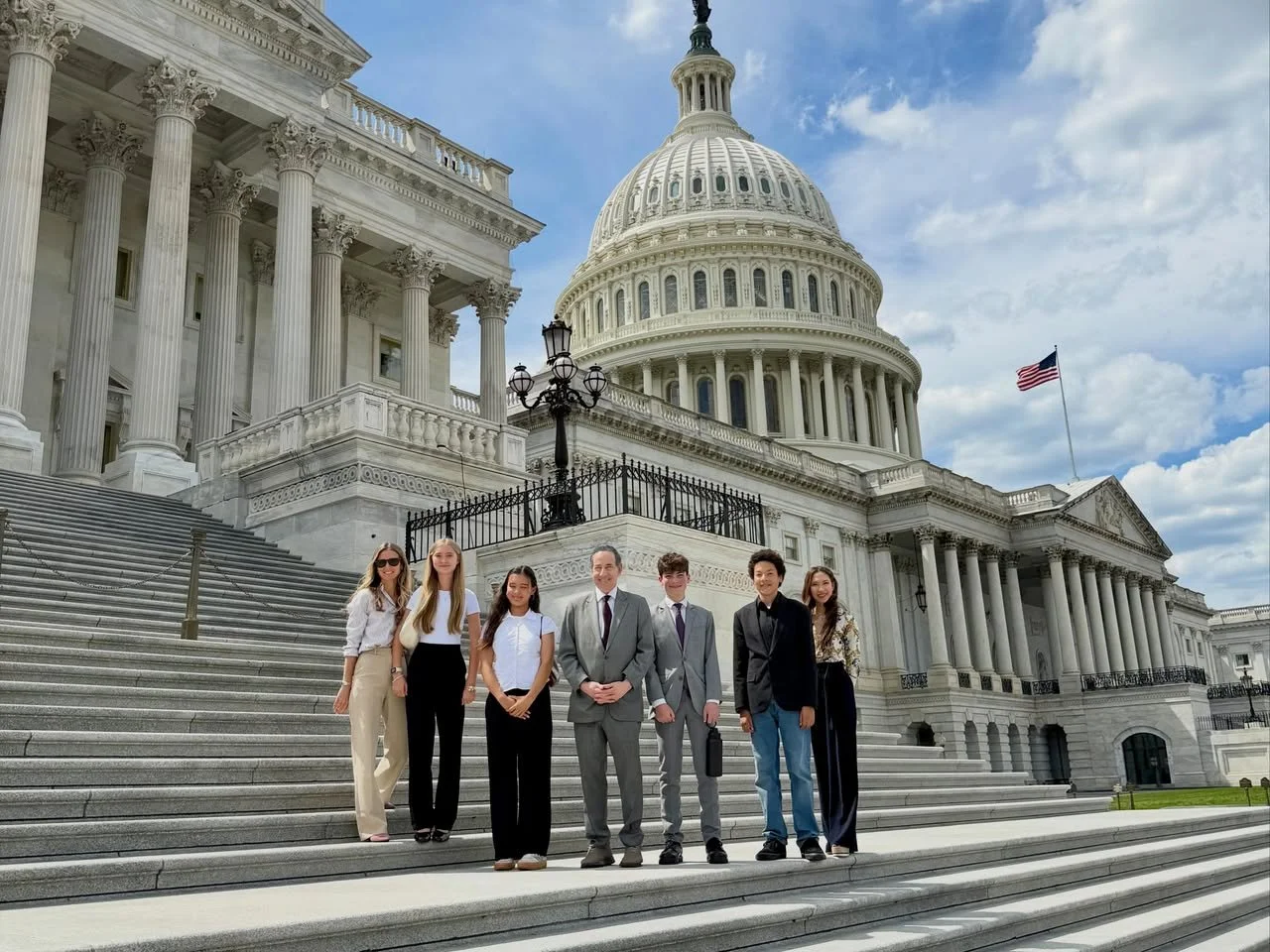 Group of seven people in business casual and formal attire standing on the steps of the U.S. Capitol building, with the dome and American flag in the background on a partly cloudy day.