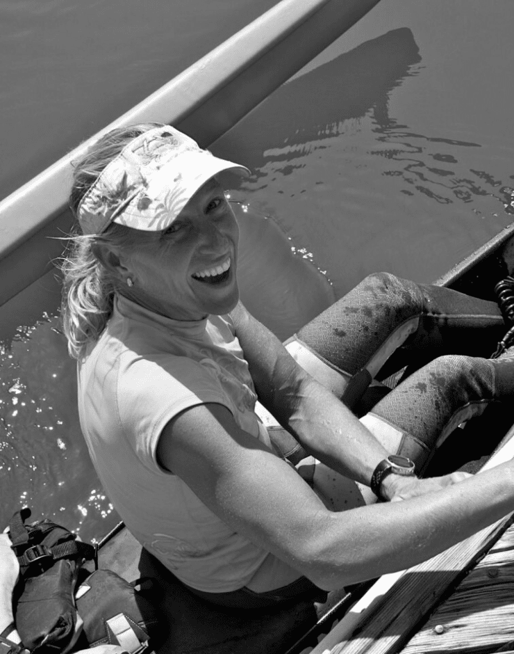 A woman sitting in a boat, smiling, wearing a visor and athletic clothes, with water around her.