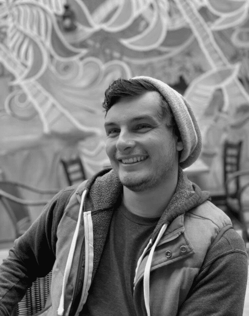 A smiling man wearing a beanie, hoodie, and jacket, sitting indoors with a graffiti mural in the background.