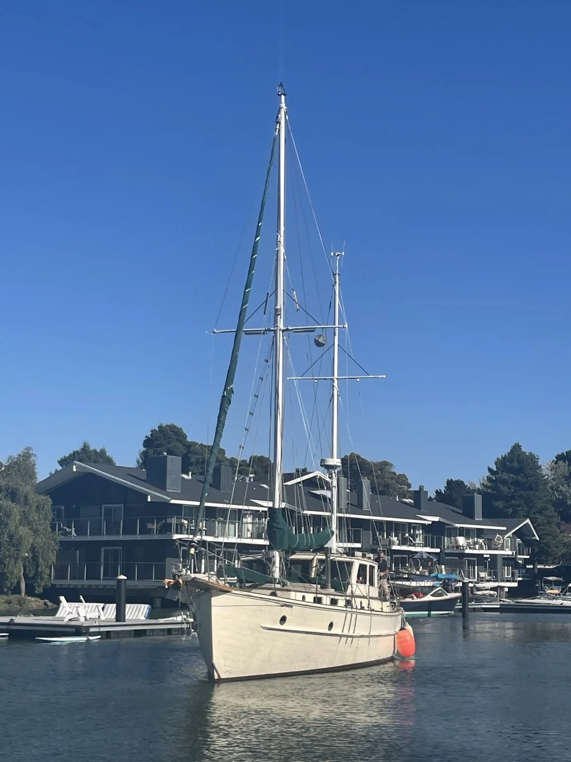 A sailboat docked near a residential building with multiple levels, surrounded by trees and a clear blue sky.