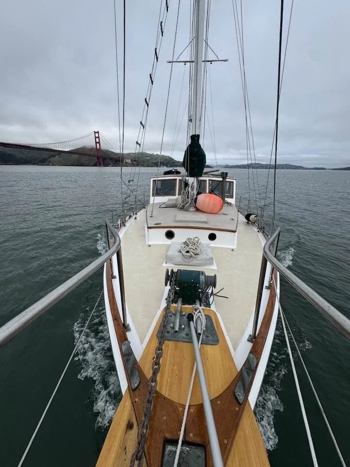 View from the bow of a sailboat with a bridge in the background, cloudy sky, and sea surrounding the boat.