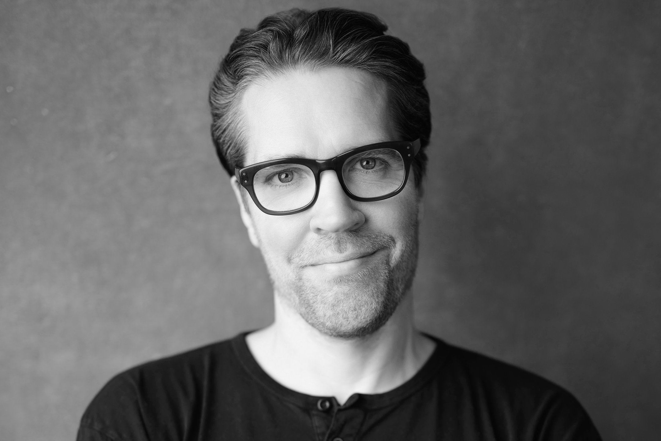 Black and white portrait of a man with glasses, short hair, and light facial hair, wearing a dark shirt, smiling slightly, against a plain background.