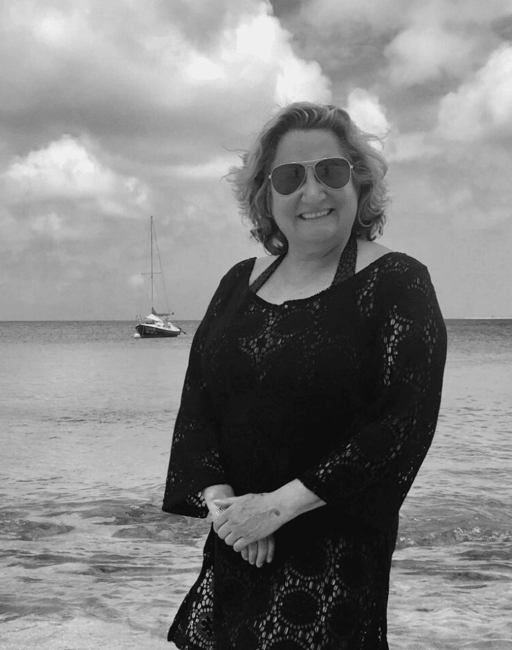 A woman smiling on a beach, wearing sunglasses and a black lace dress, with a boat in the water and cloudy sky in the background.