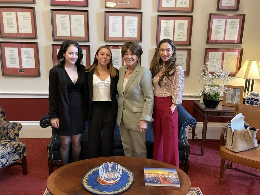 Four women standing together in a formal room with framed documents on the wall behind them. Two young women on each side of an older woman in the middle. The woman on the far right is wearing a patterned blouse and red wide-legged pants, while the w