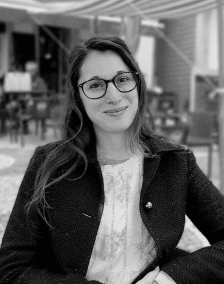 A woman wearing glasses and a coat smiling at the camera in a room with wooden walls and furniture.