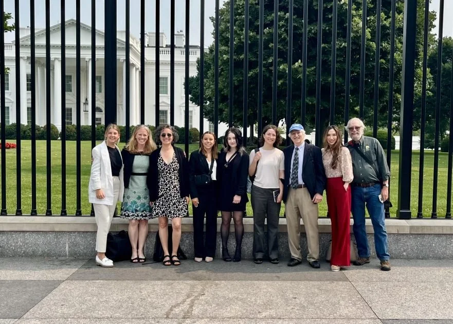 Group of nine people standing in front of a black fence with the White House in the background.