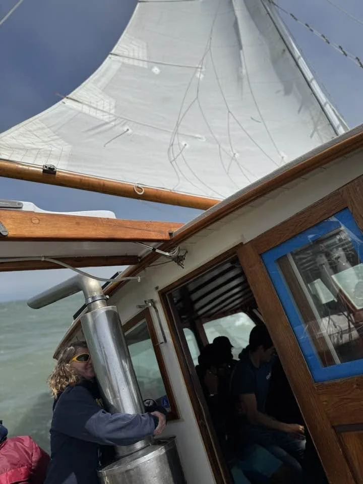 View from a sailboat showing the sail, the boat's wooden structure, and some people inside and on deck.