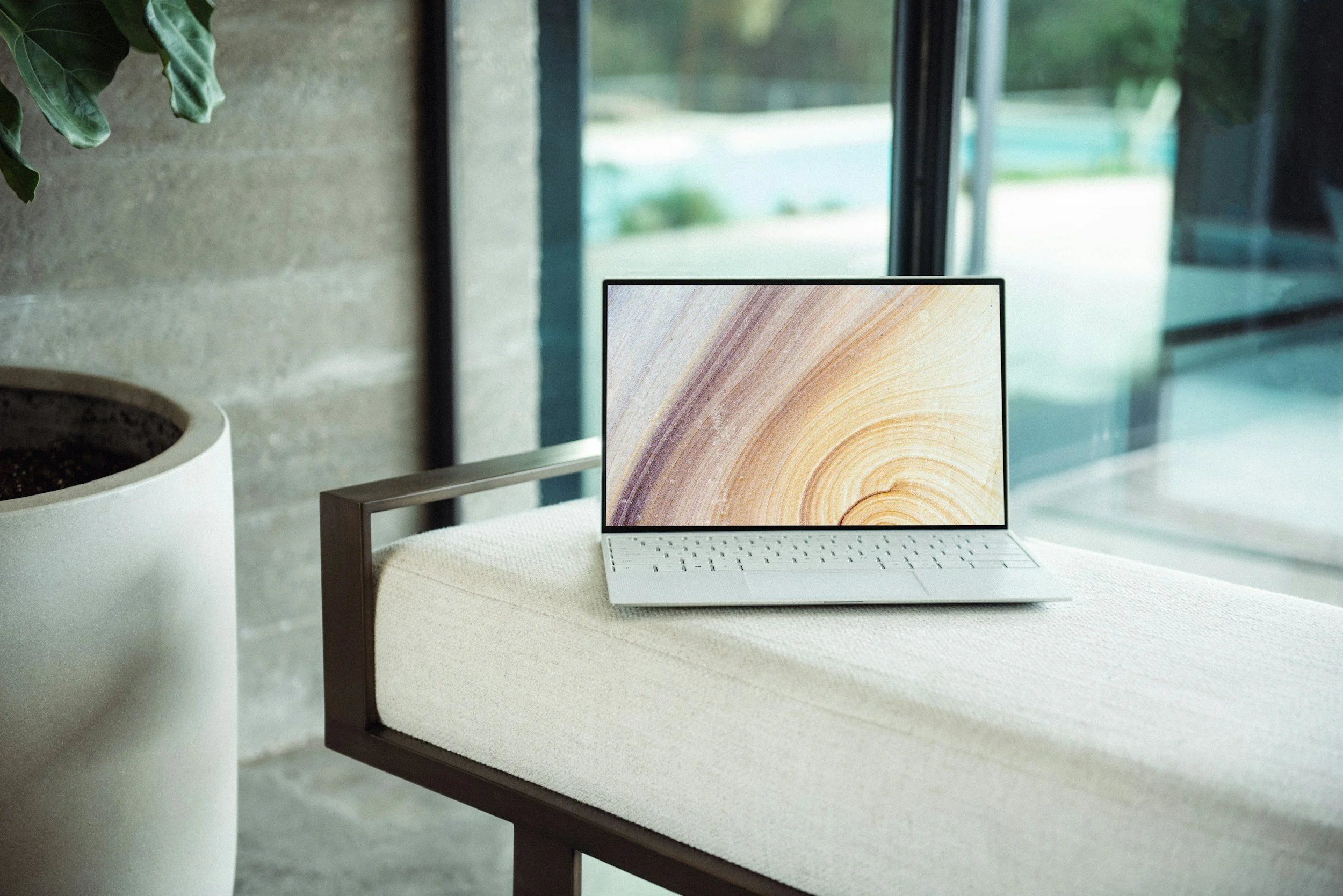 A slim white laptop with a wooden swirl pattern on the screen, placed on a beige cushioned chair next to a white cylindrical pot with green foliage, in front of a large glass window showing an outdoor scene with greenery.