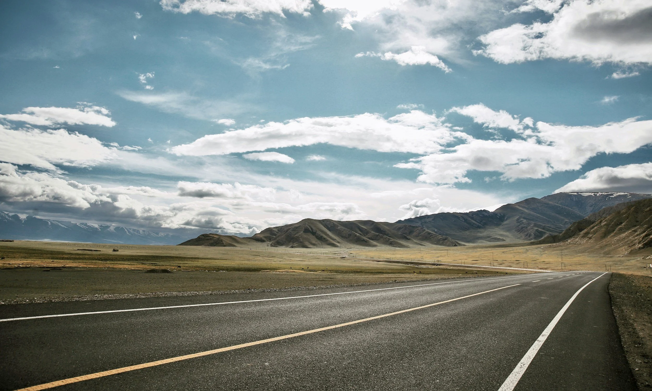 A long, empty asphalt highway stretches through a wide valley with grassy plains and distant majestic mountains under a partly cloudy sky and a luminous sun.