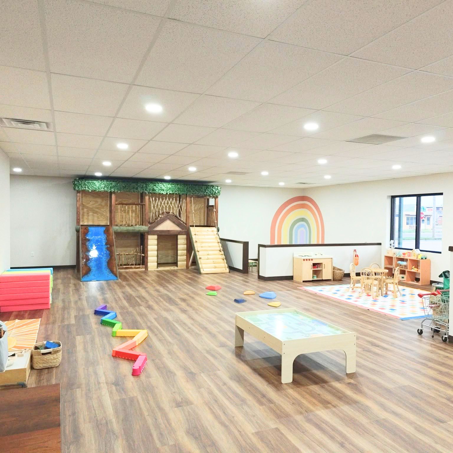 Bright and spacious indoor classroom with wooden flooring, playful climbing structures, and small furniture for children. There is a rainbow mural on the wall, a rug with a colorful pattern, and large windows letting in natural light.