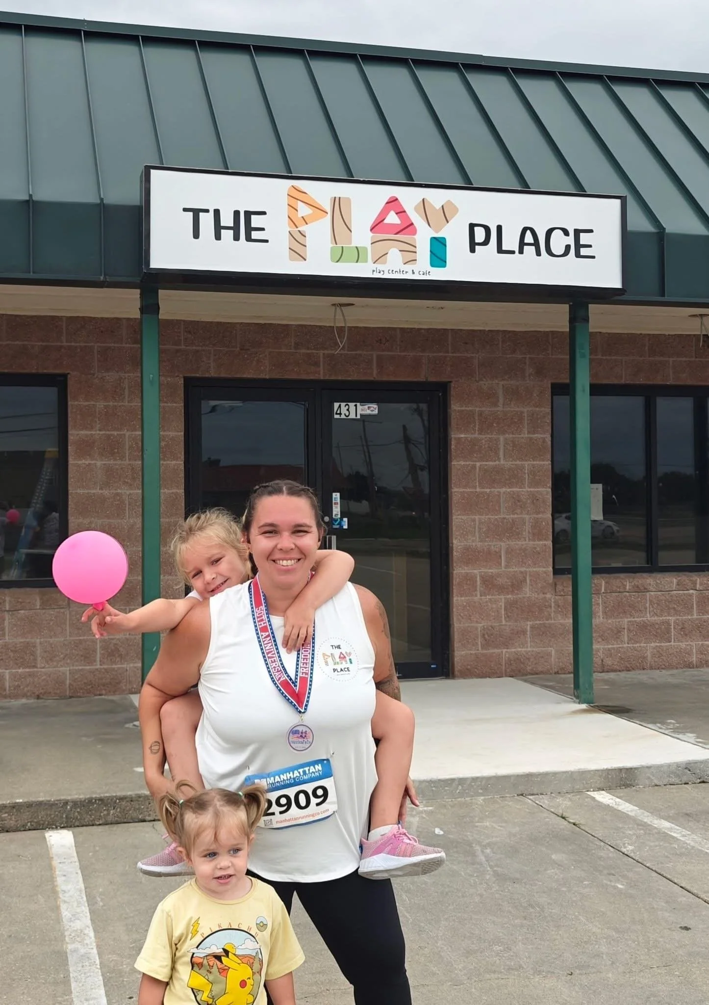 A woman holding a young girl on her shoulders, both smiling, standing outside a building with a sign that says 'The Play Place,' and two small girls in front of them, one with a pink balloon. The woman is wearing a medal and a race bib, indicating participation in a race or marathon.