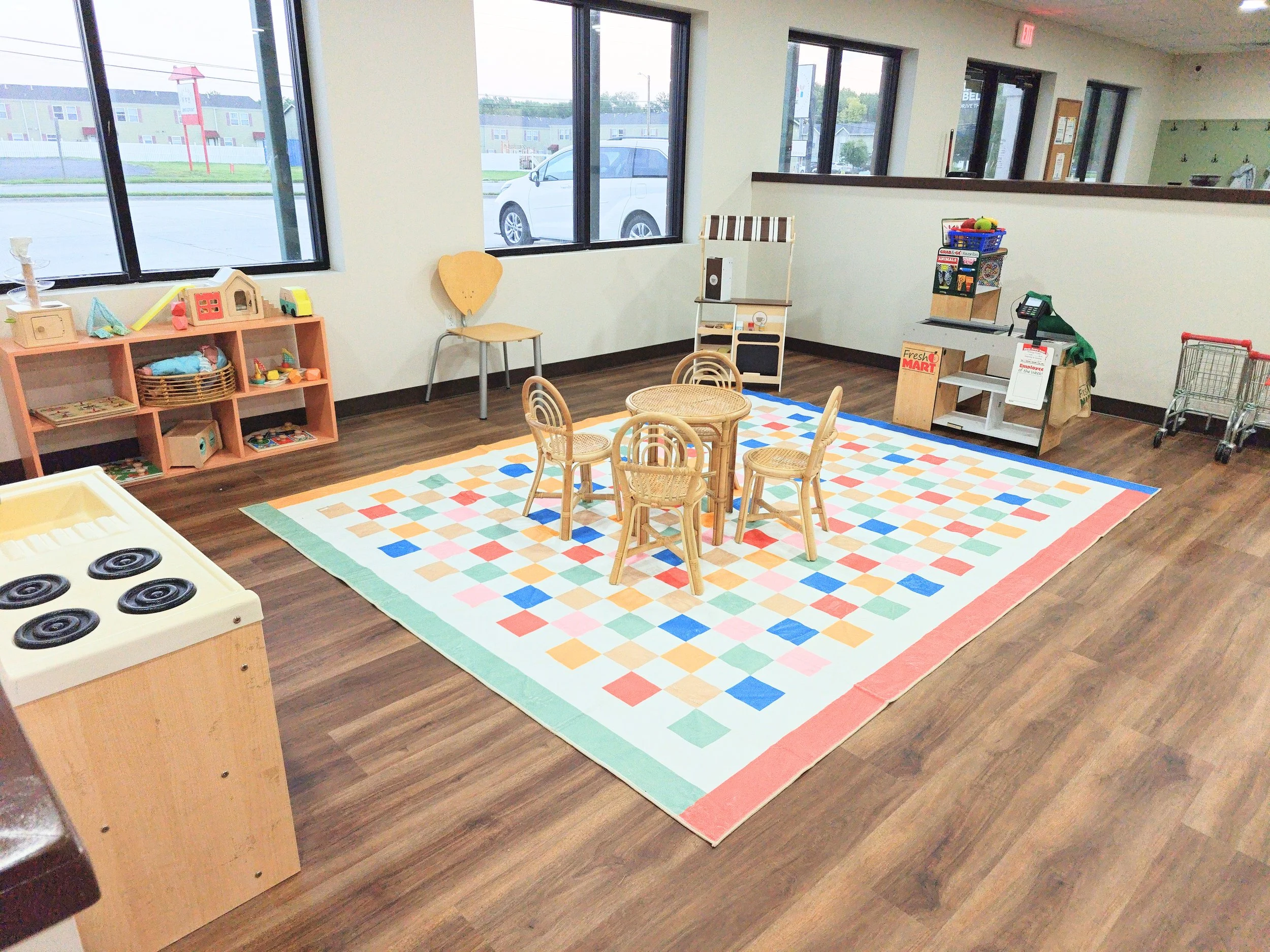 A children's play area with wooden furniture, a colorful checkered rug, toys, a small shopping cart, and large windows in a room with wooden flooring.