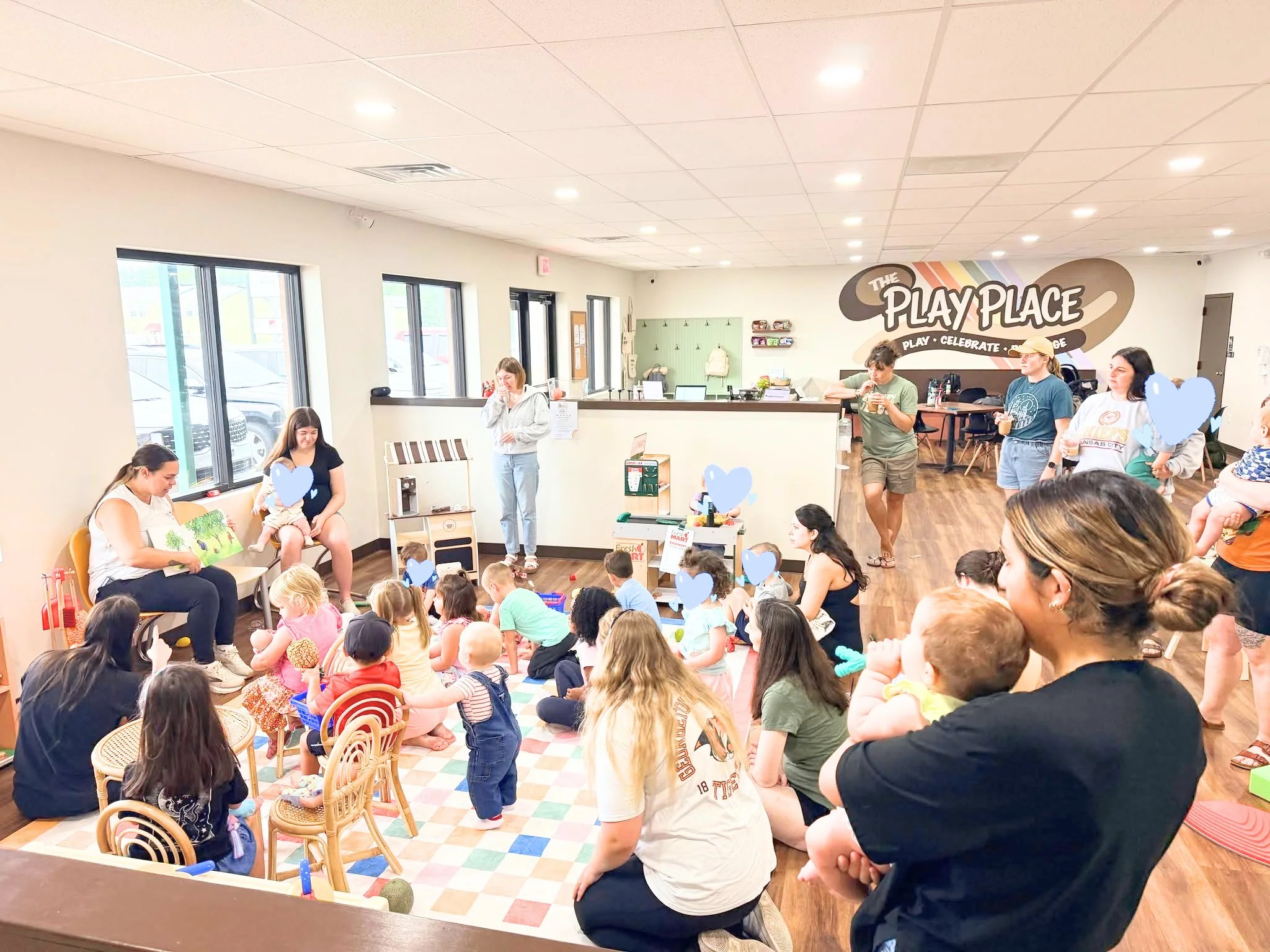 People gathered in a community room with children sitting on the floor, some holding balloons and toys, and adults watching a woman reading a book, with a wall sign that says 'The Play Place'.