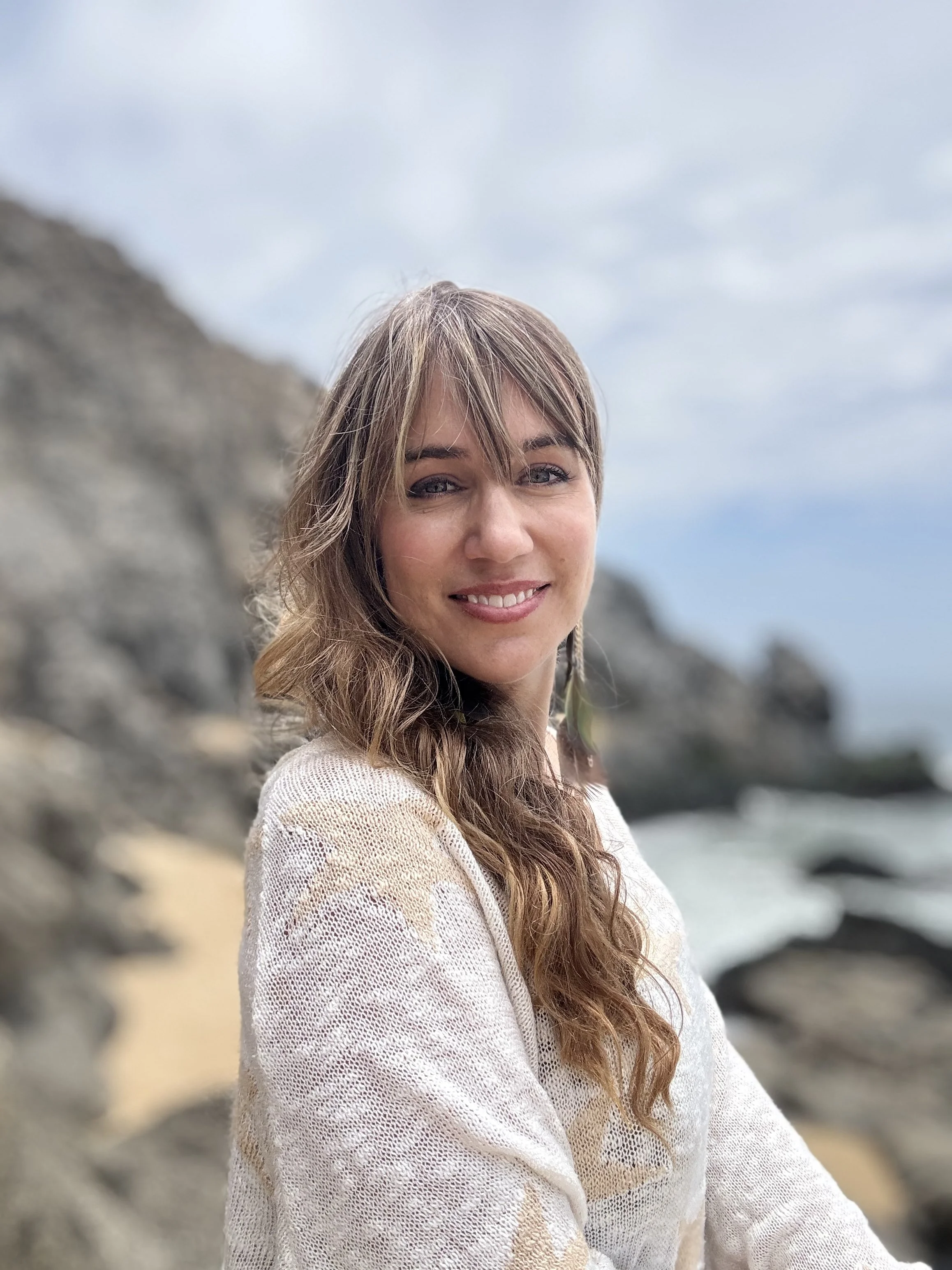 A woman with long, wavy hair smiling at the camera outdoors, with rocky terrain and a cloudy sky in the background.
