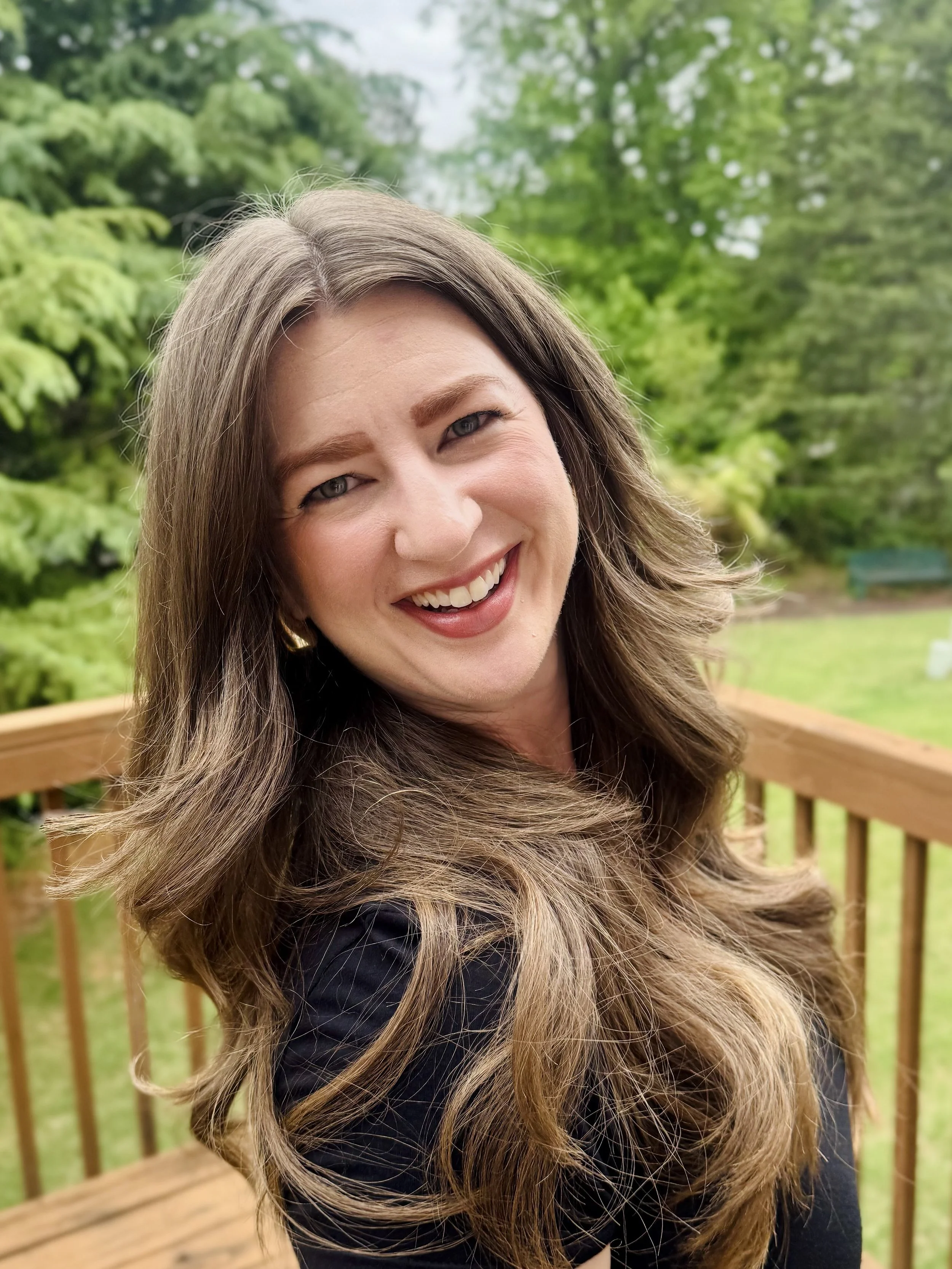 A woman with long, wavy brown hair and a bright smile standing outdoors on a wooden deck with lush green trees in the background.