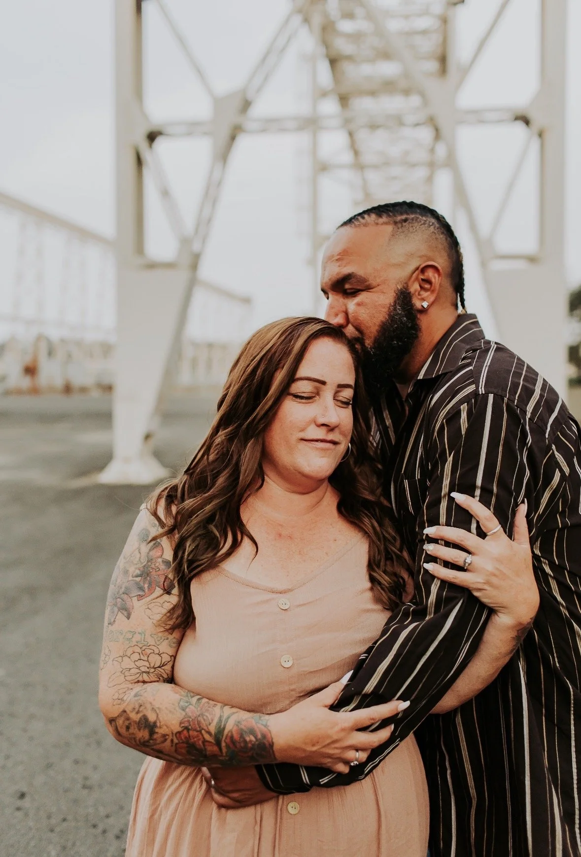 A man with a beard and earrings is giving a kiss on the forehead to a woman with tattoos on her arms, both embracing each other outside near a large industrial structure.