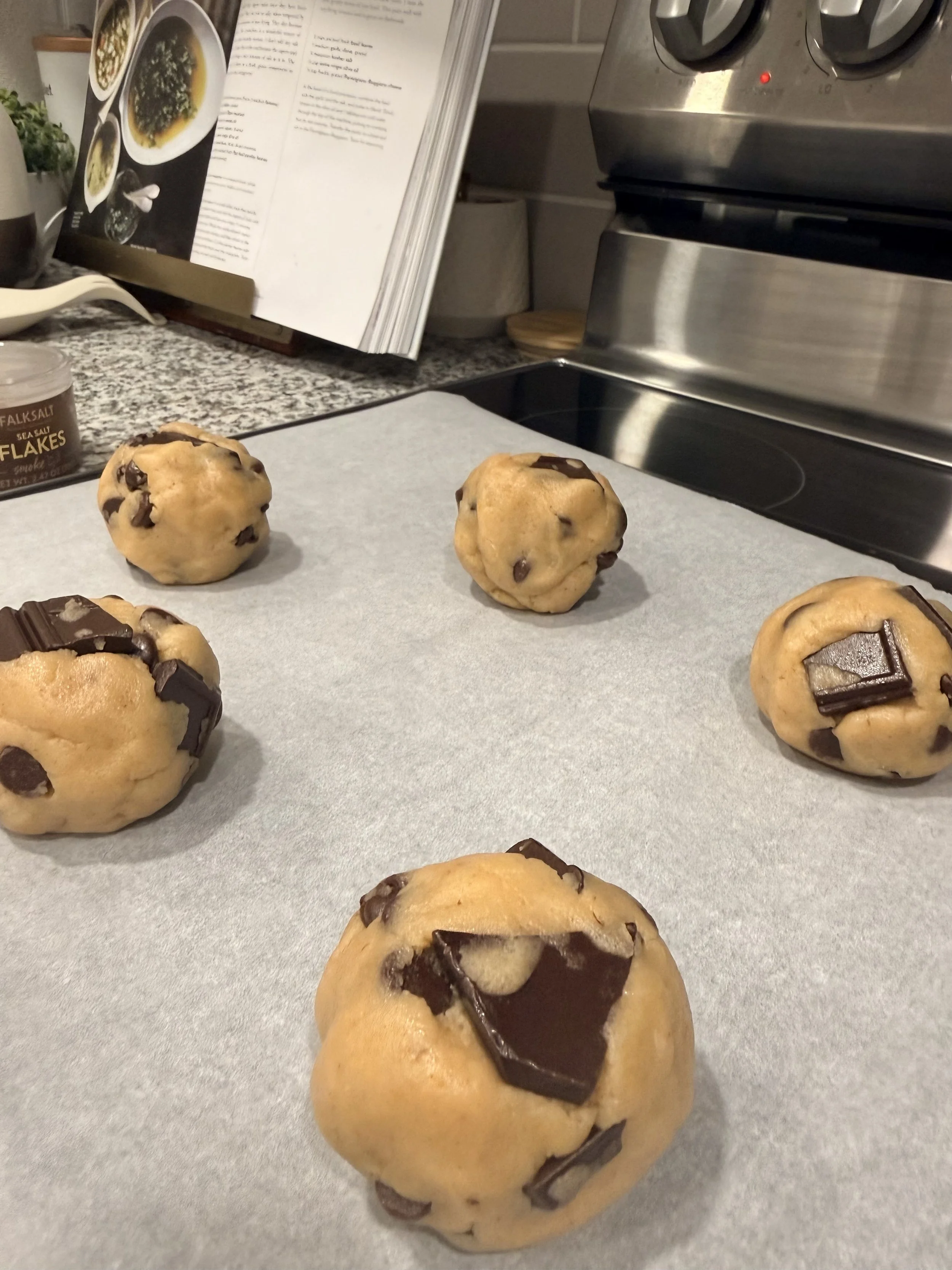 Chocolate chip cookies dough on a baking sheet in a kitchen, with a cookbook or recipe book open in the background.