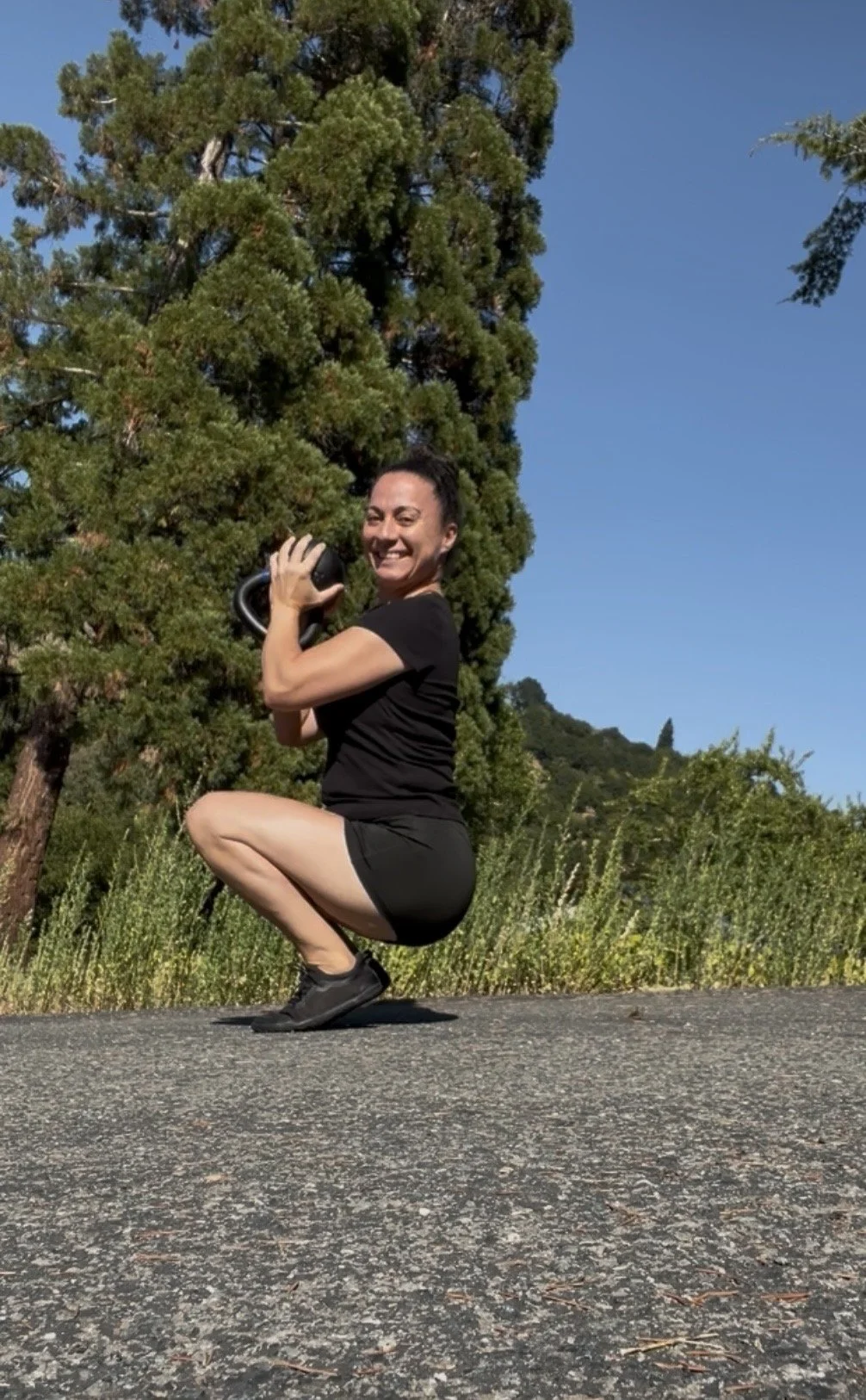 A woman smiling while holding a kettlebell outdoors on a paved path with green trees in the background.