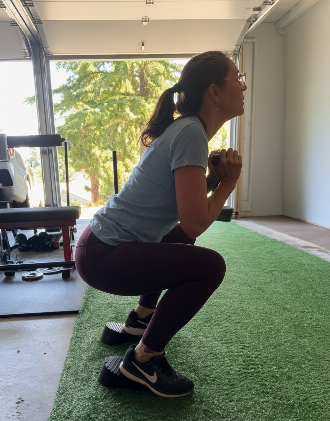 Woman in workout clothes squatting while holding a dumbbell in a gym with a garage door open and green trees outside.